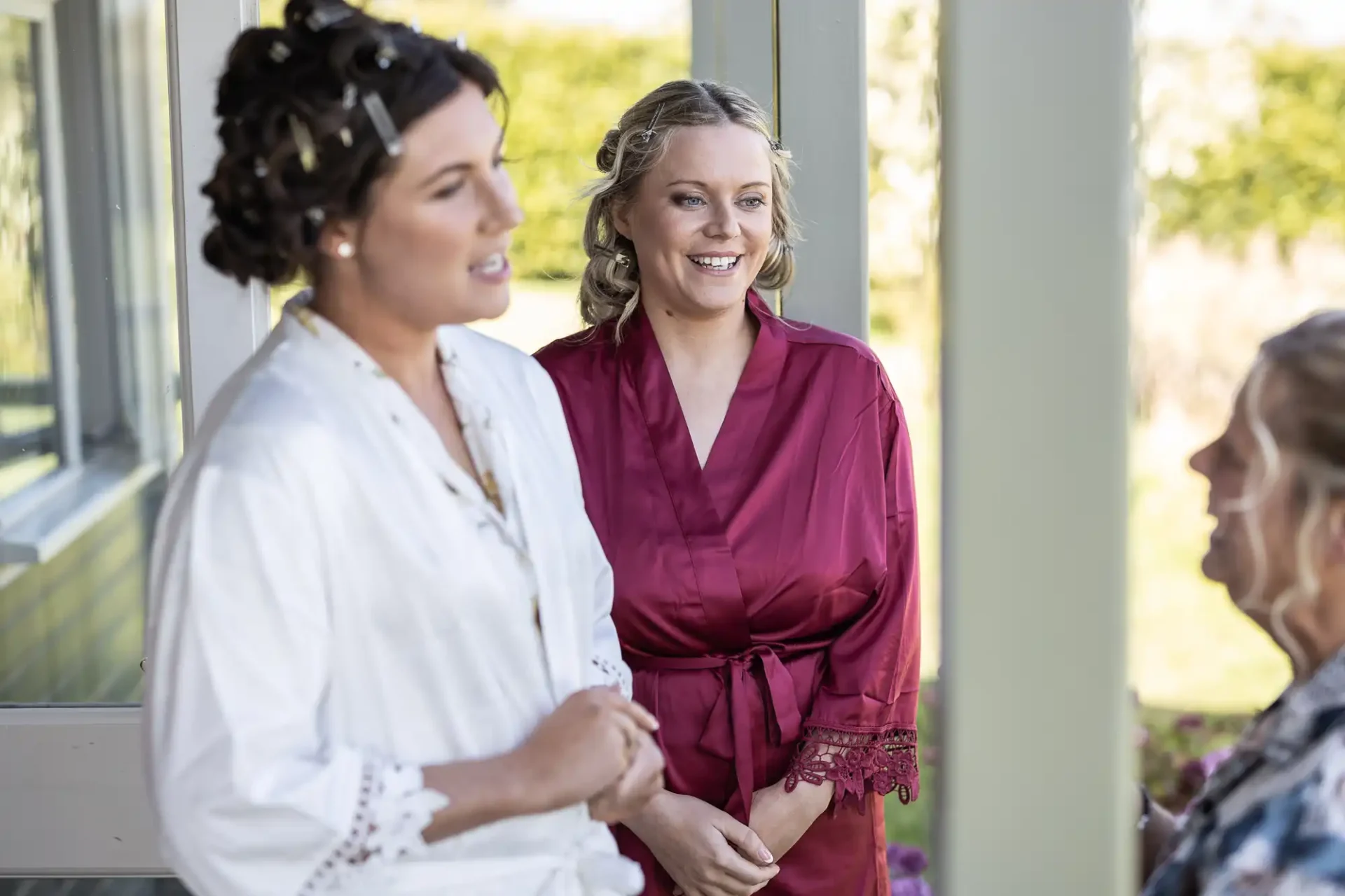 Two women in robes, one in white, the other in red, stand talking with a third person partially visible. They are outdoors next to a building with greenery in the background.