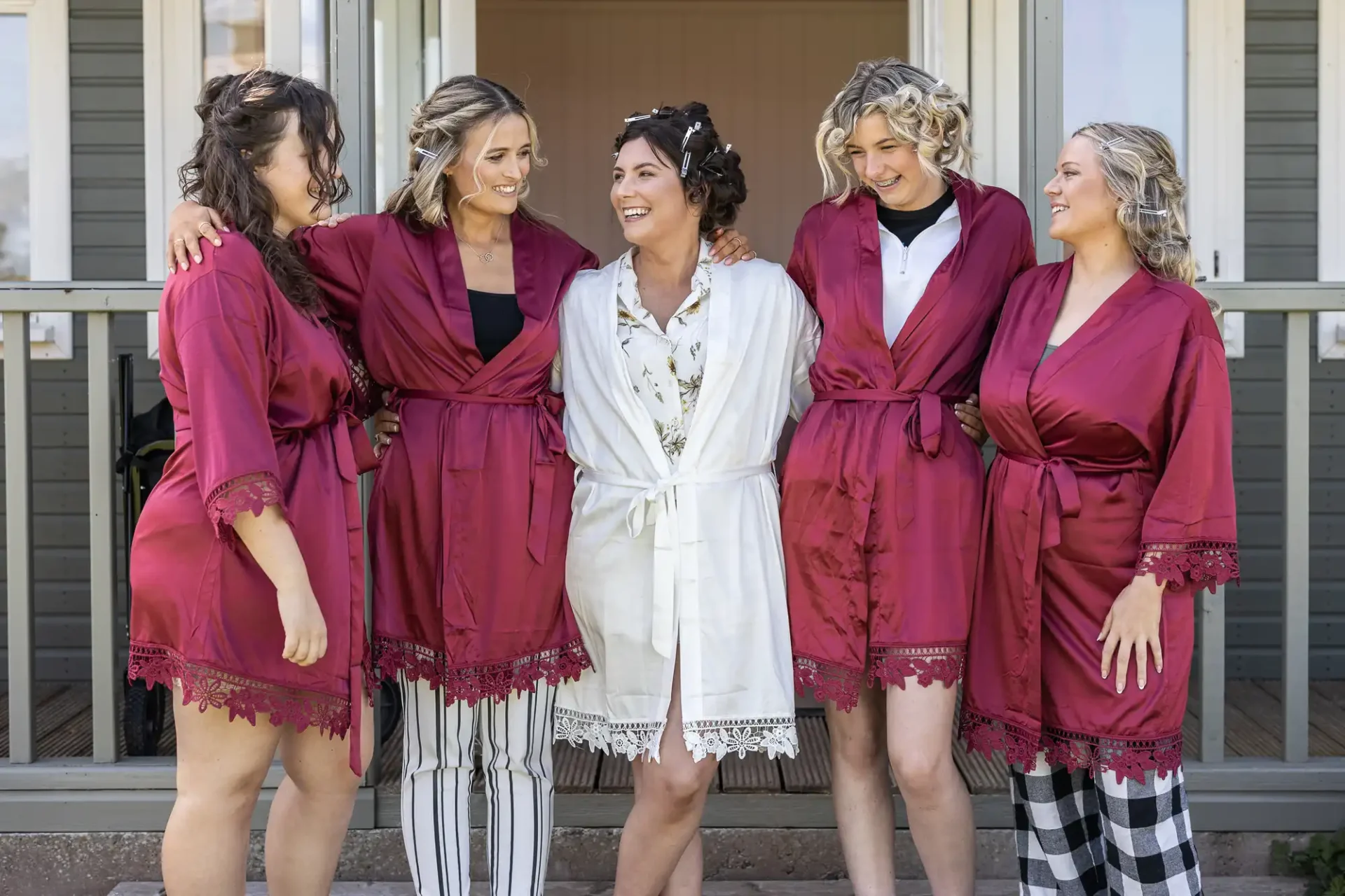 Five women standing together, one in a white robe and the others in matching red robes and pajamas, in front of a building with columns.