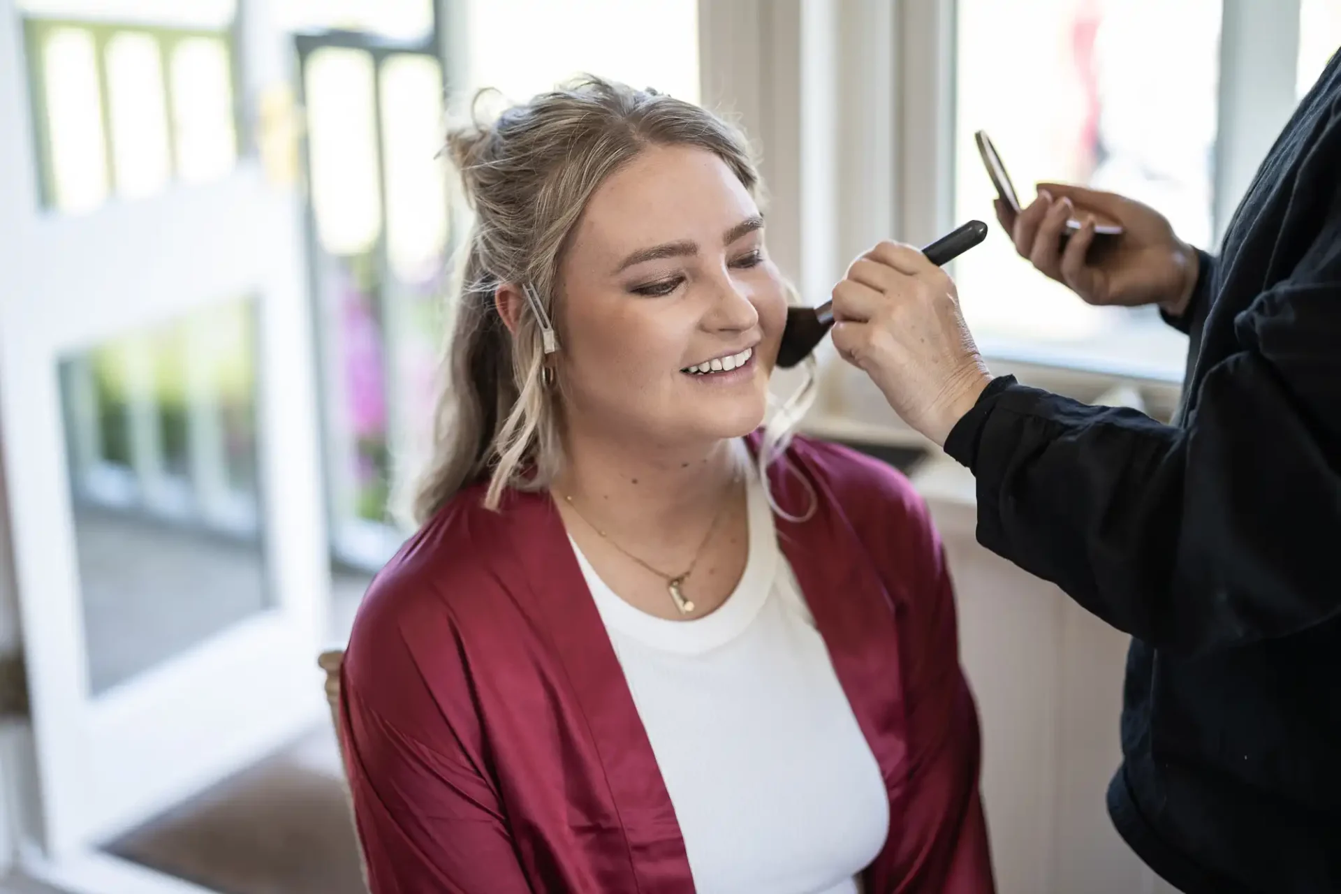 A woman in a red robe sits indoors while a person applies makeup to her face with a brush.