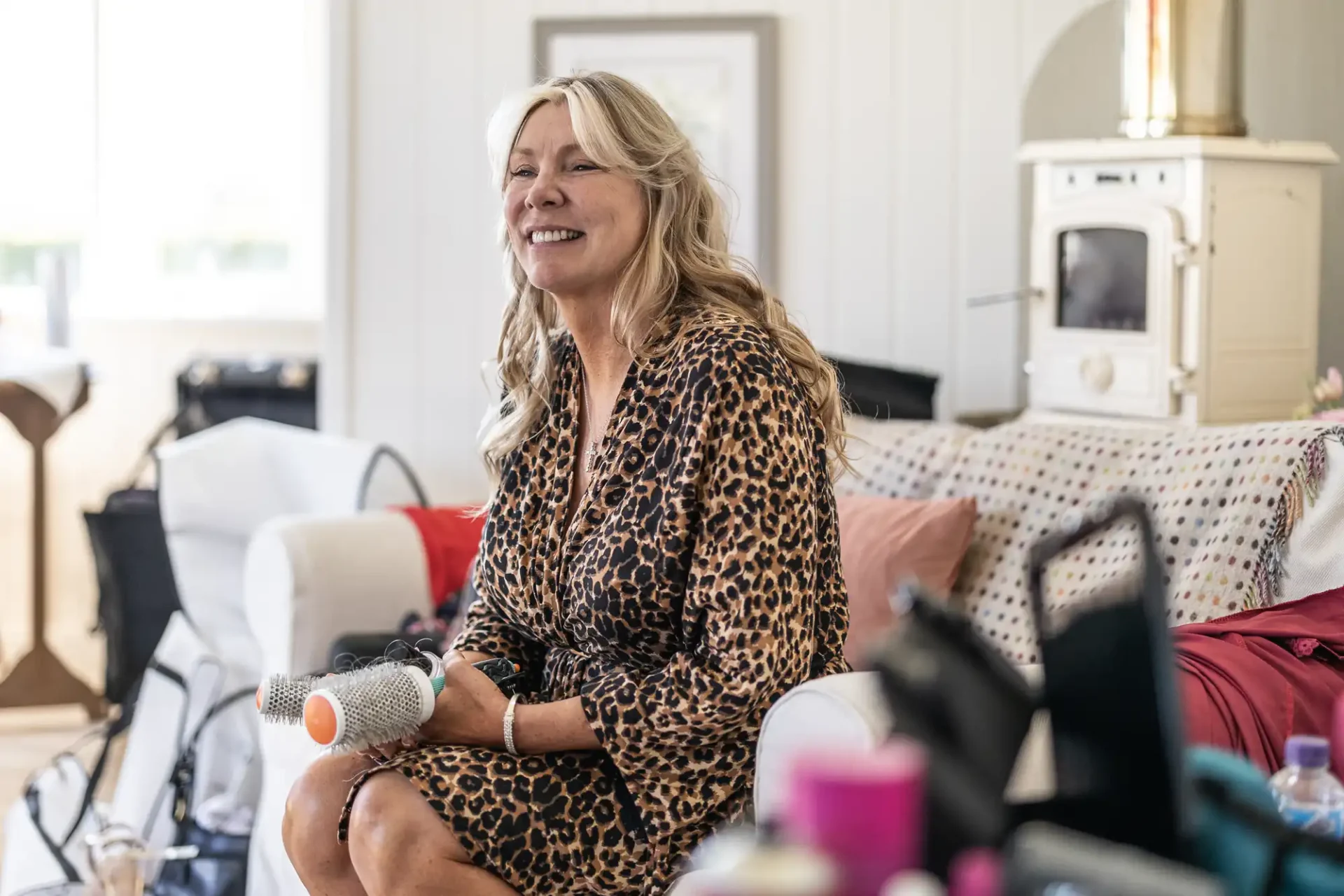 Woman in a leopard print outfit sitting on a couch, holding a round hairbrush, smiling. Background includes a vintage oven and various items.