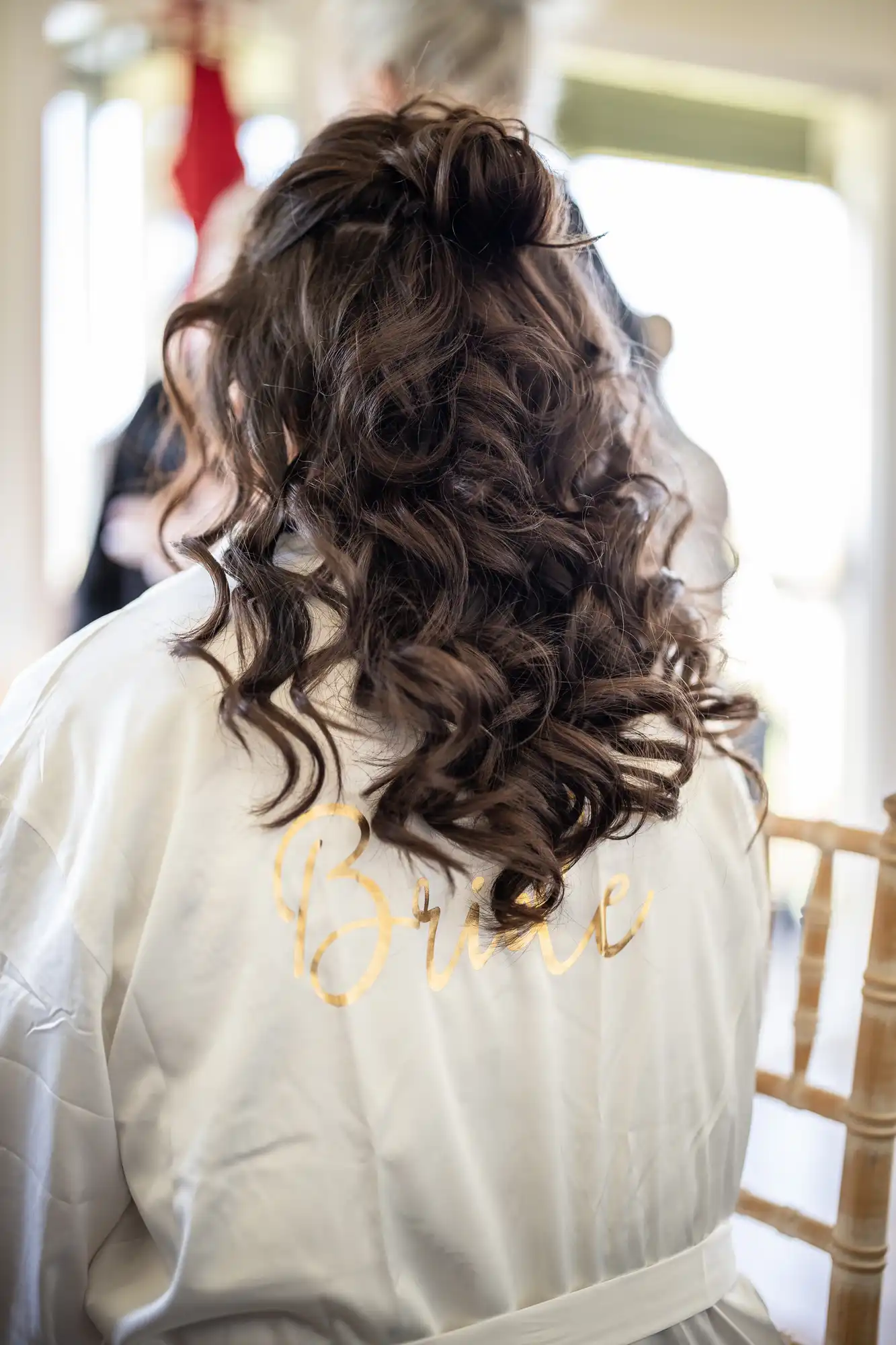 A woman with long, curly brown hair is seen from behind, wearing a white robe with "Bride" written on it in gold lettering. She is seated indoors.