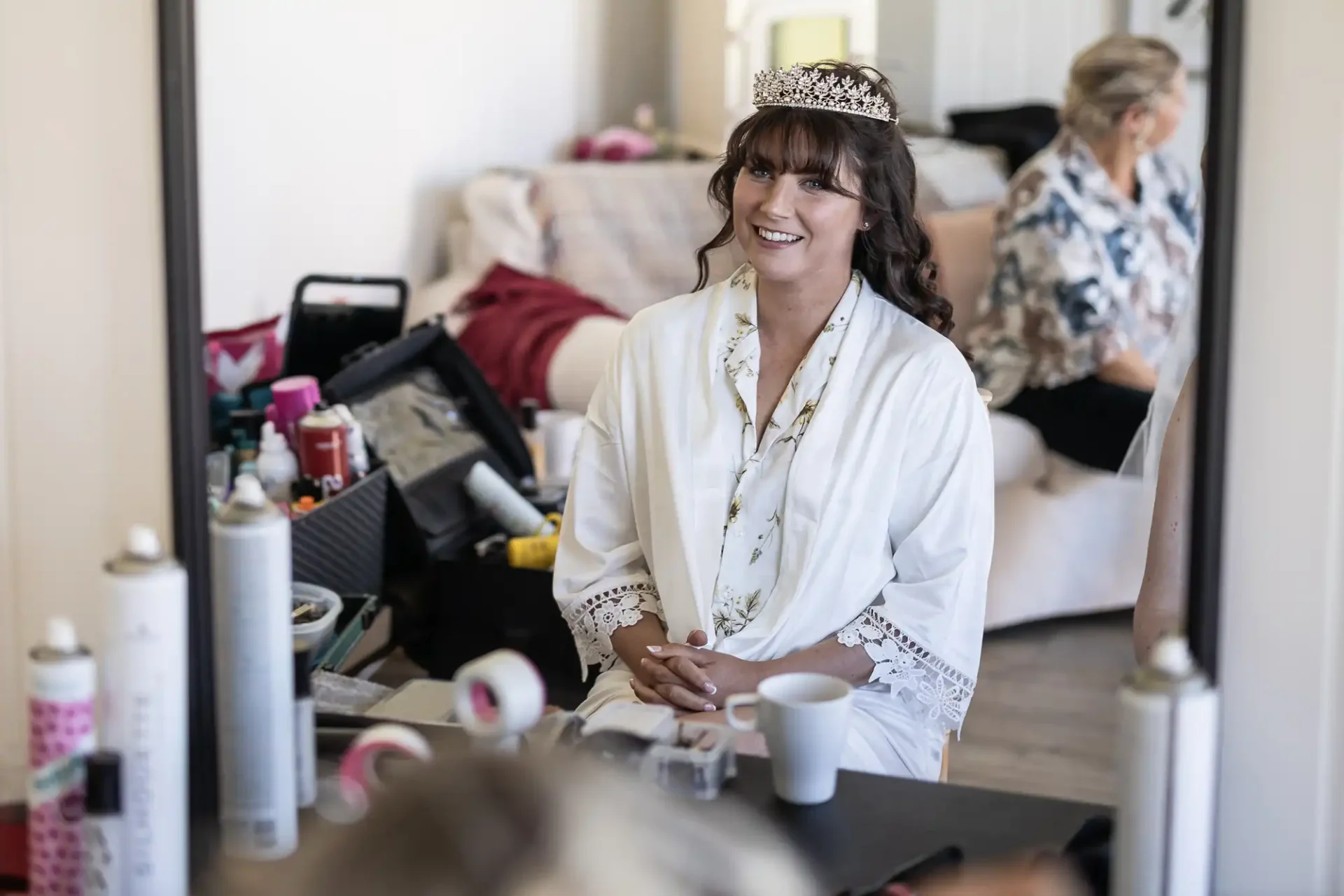 A woman in a white robe and a tiara sits smiling in front of a mirror, surrounded by cosmetics and styling tools. Another person is seated in the background.