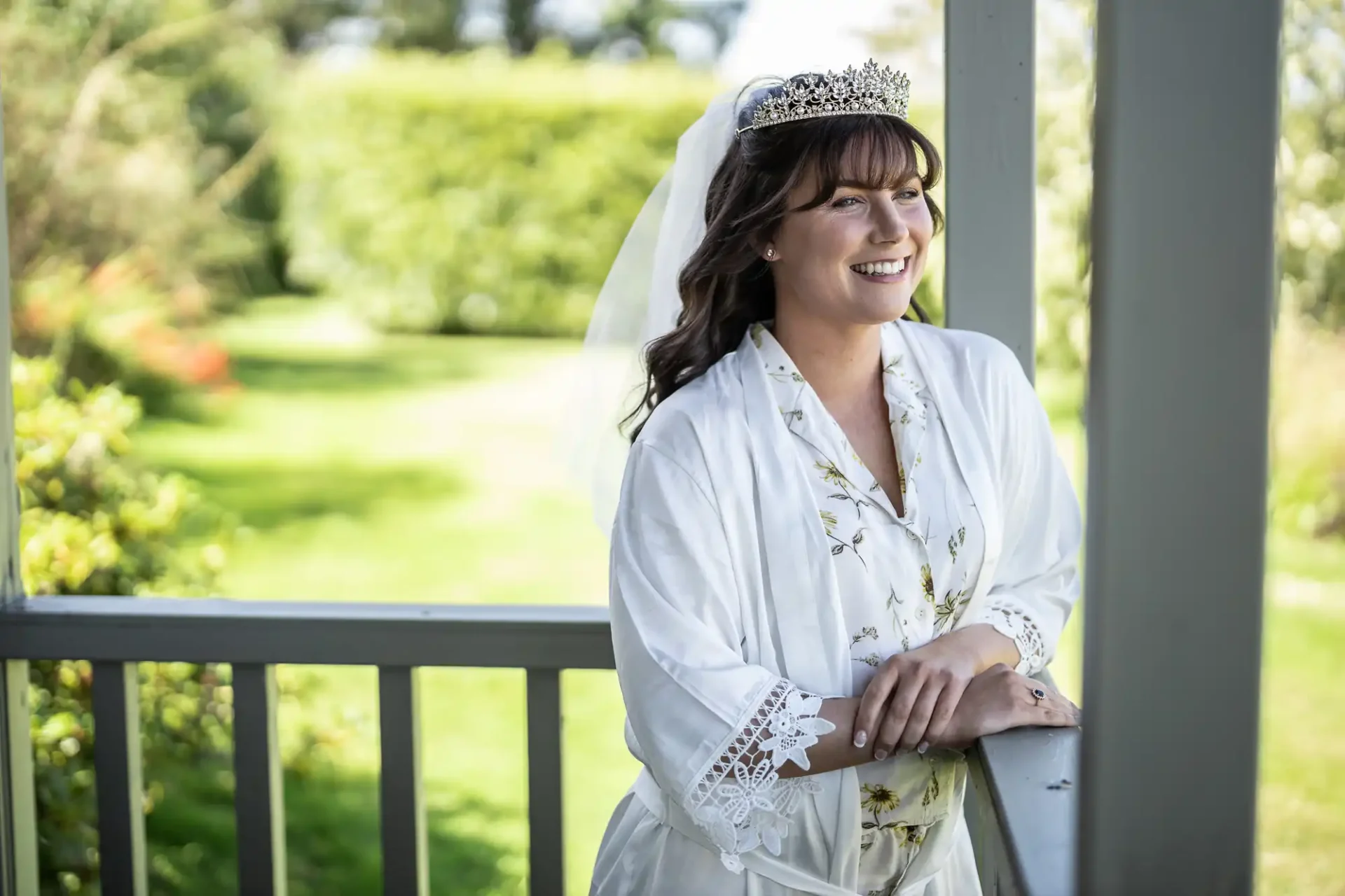 A woman in a white robe and tiara stands on a porch, smiling with greenery in the background.