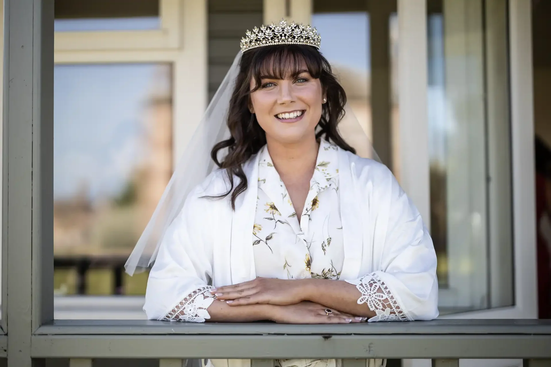 A smiling bride in a white robe and tiara poses by a window, hands folded on the sill.