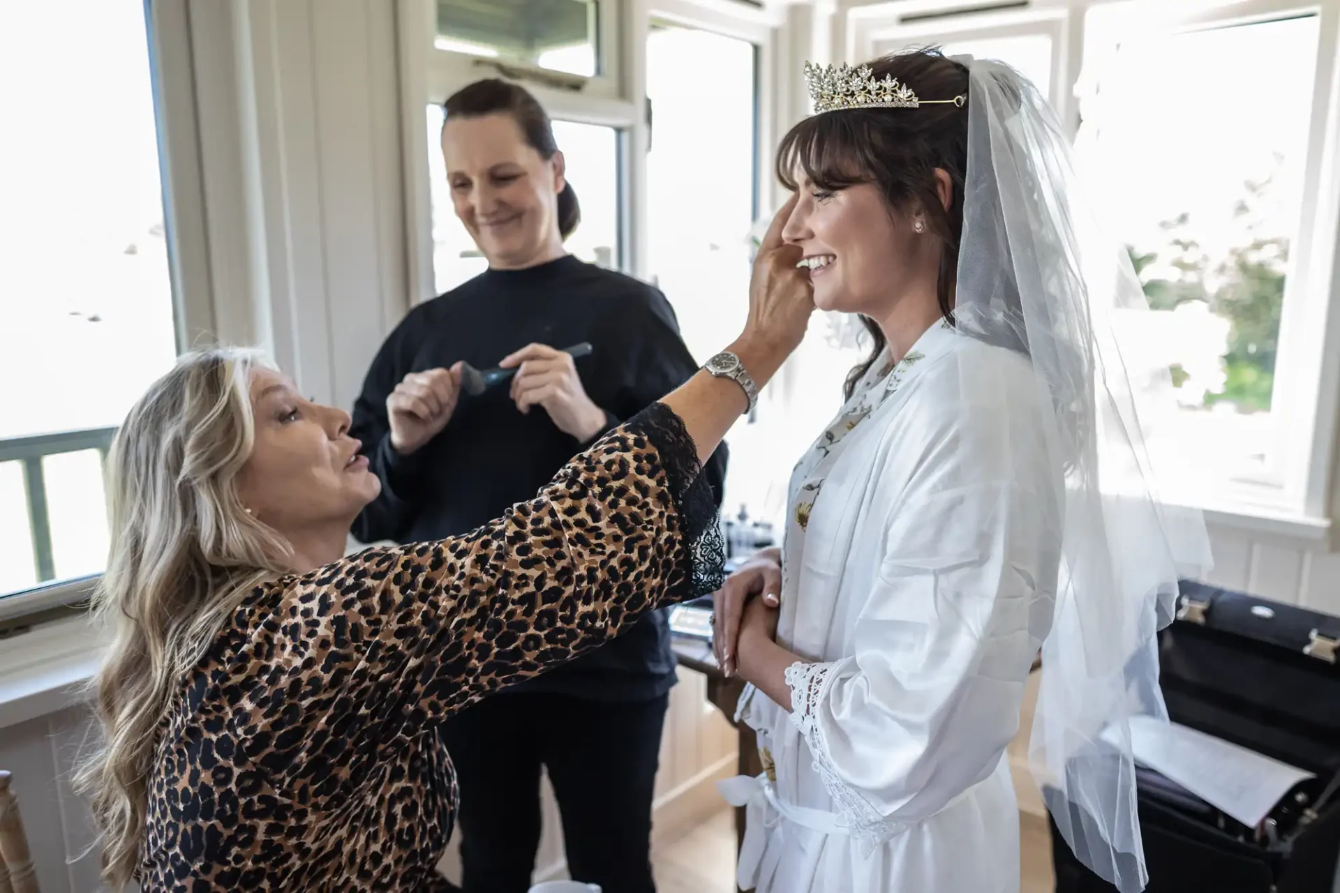 A bride in a white robe and tiara is having makeup applied by a woman, while another woman stands nearby holding a brush.