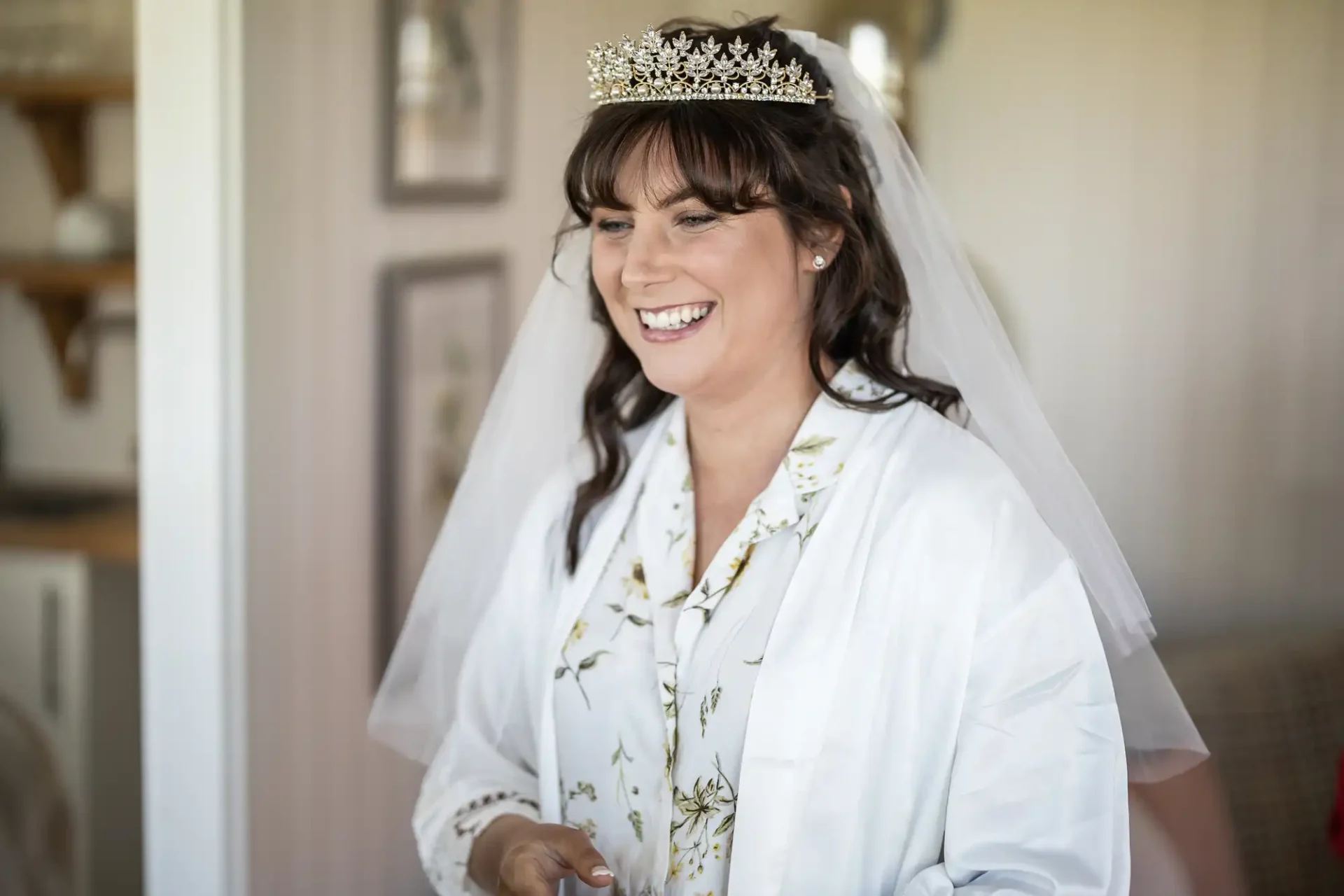 A smiling bride wearing a tiara and veil is dressed in a floral robe, standing indoors.