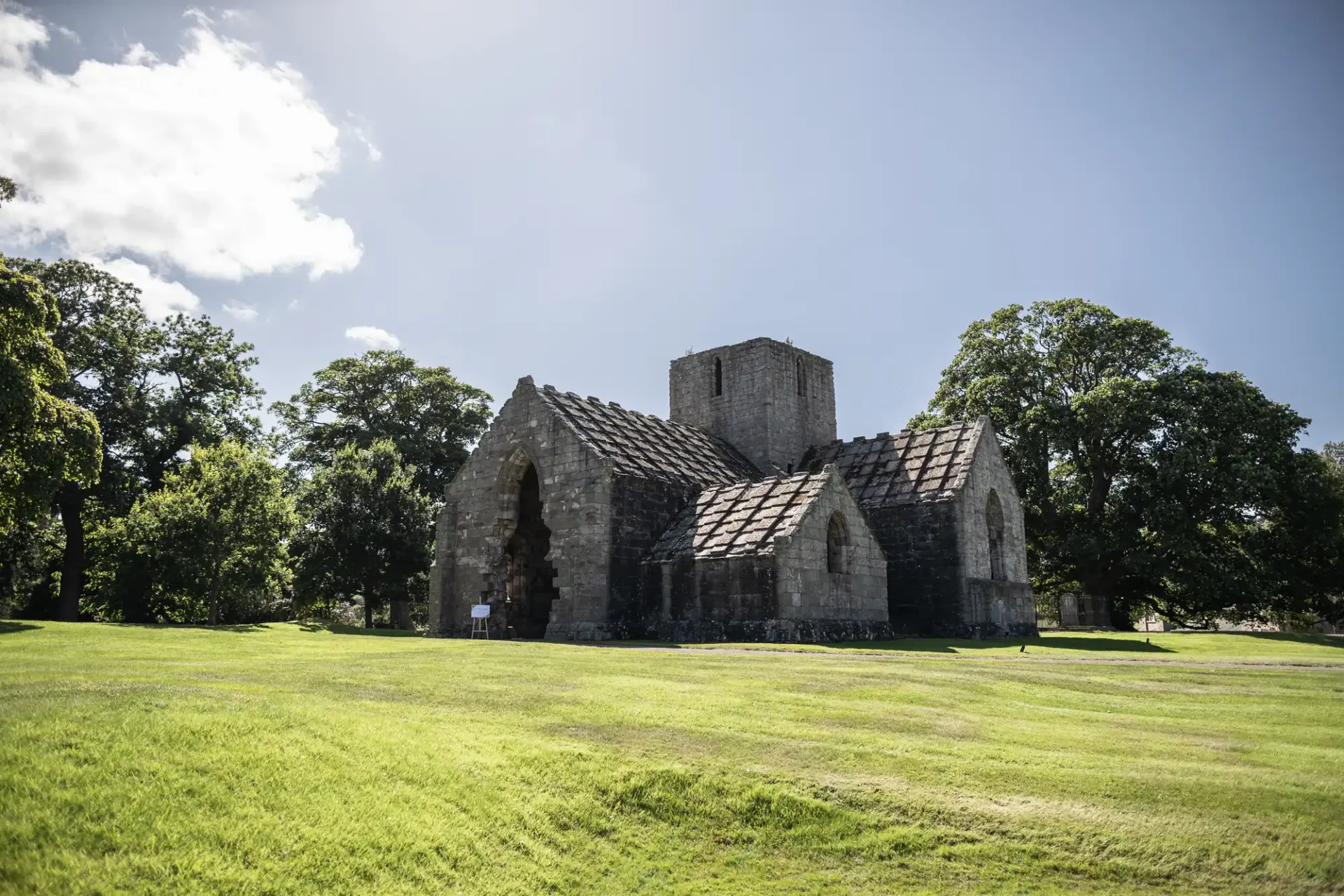 An old stone church with a central tower, surrounded by a grassy field and trees under a clear blue sky.