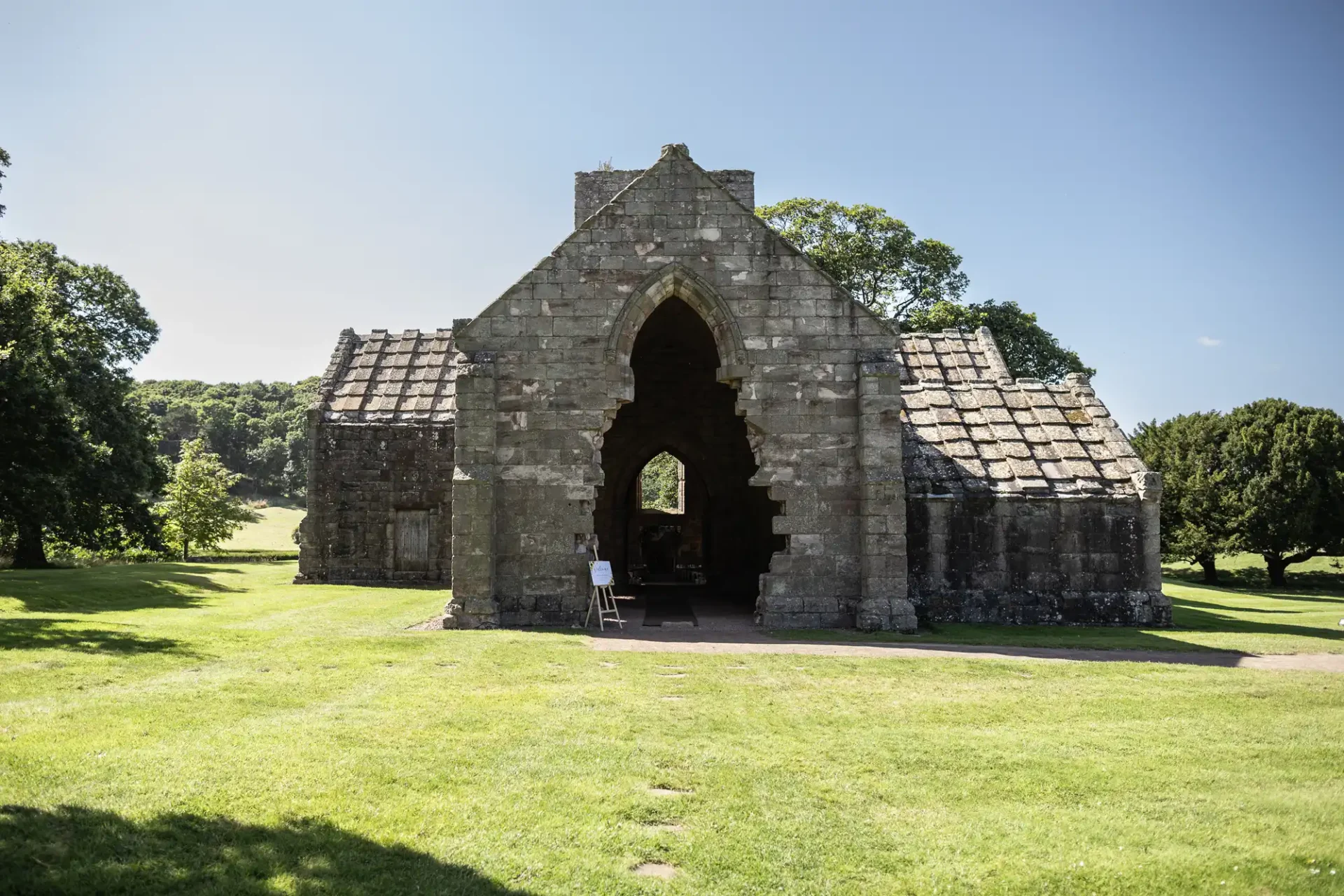 Stone building with an arched entrance in a grassy field, surrounded by trees under a clear blue sky.