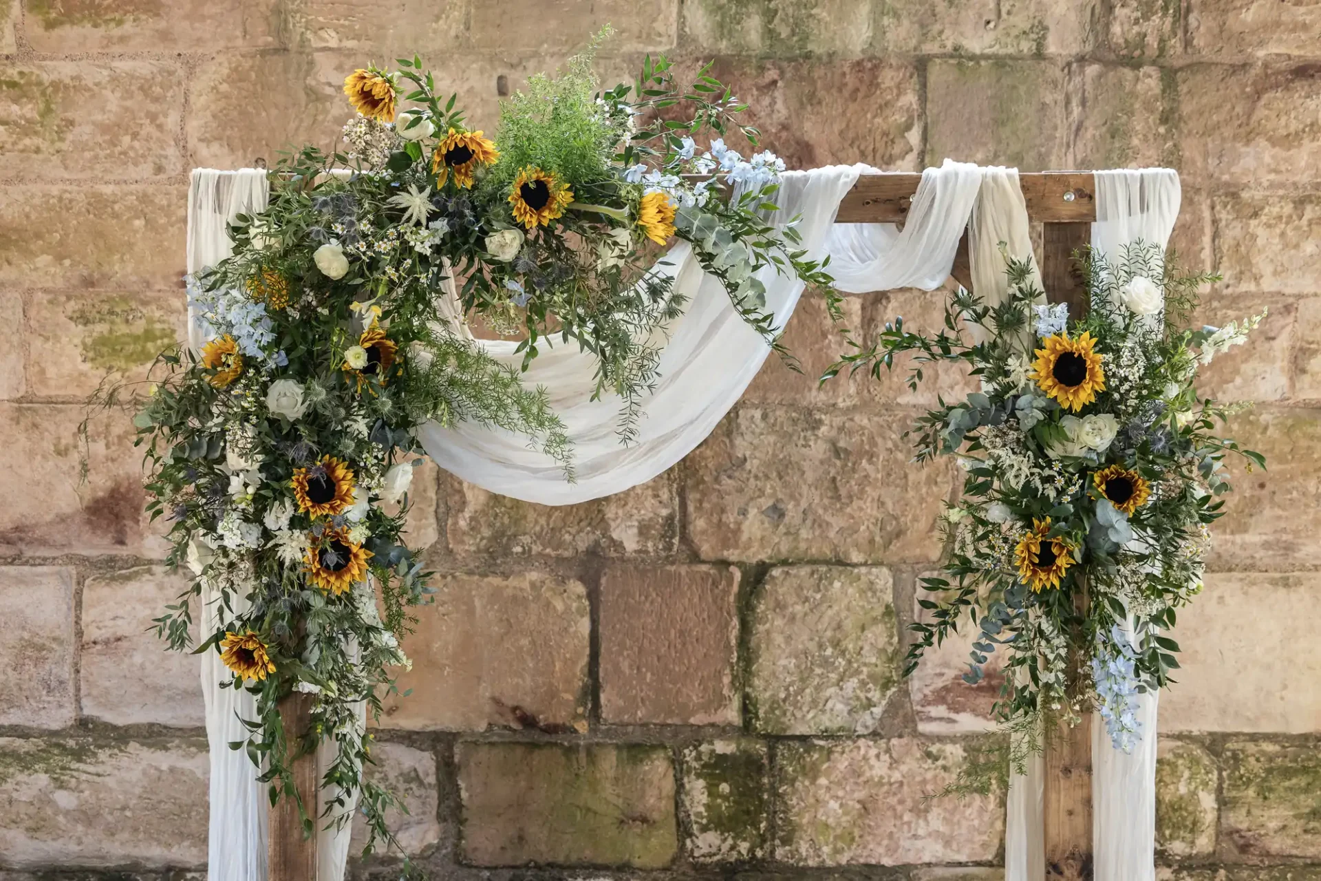 Wooden arch decorated with sunflowers, white flowers, and greenery, draped with white fabric, set against a stone wall.