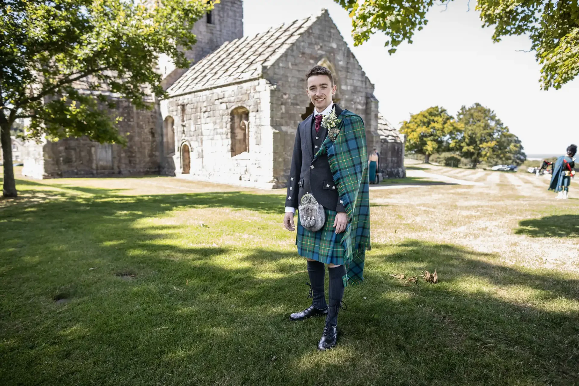 Man in a tartan kilt and dark jacket stands smiling on grass near stone building and trees.