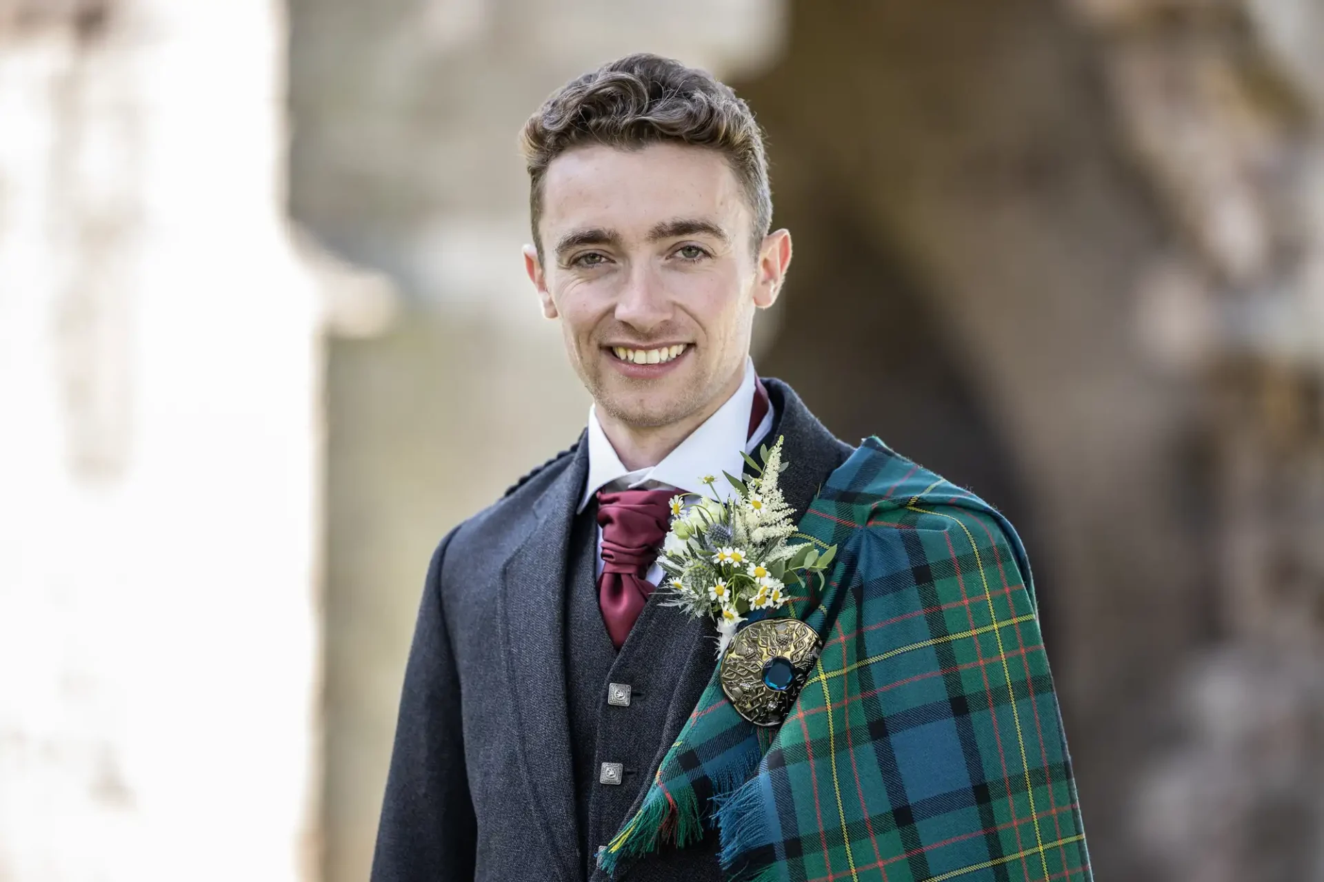 A man wearing a dark suit, red tie, and a tartan sash stands outdoors, smiling.