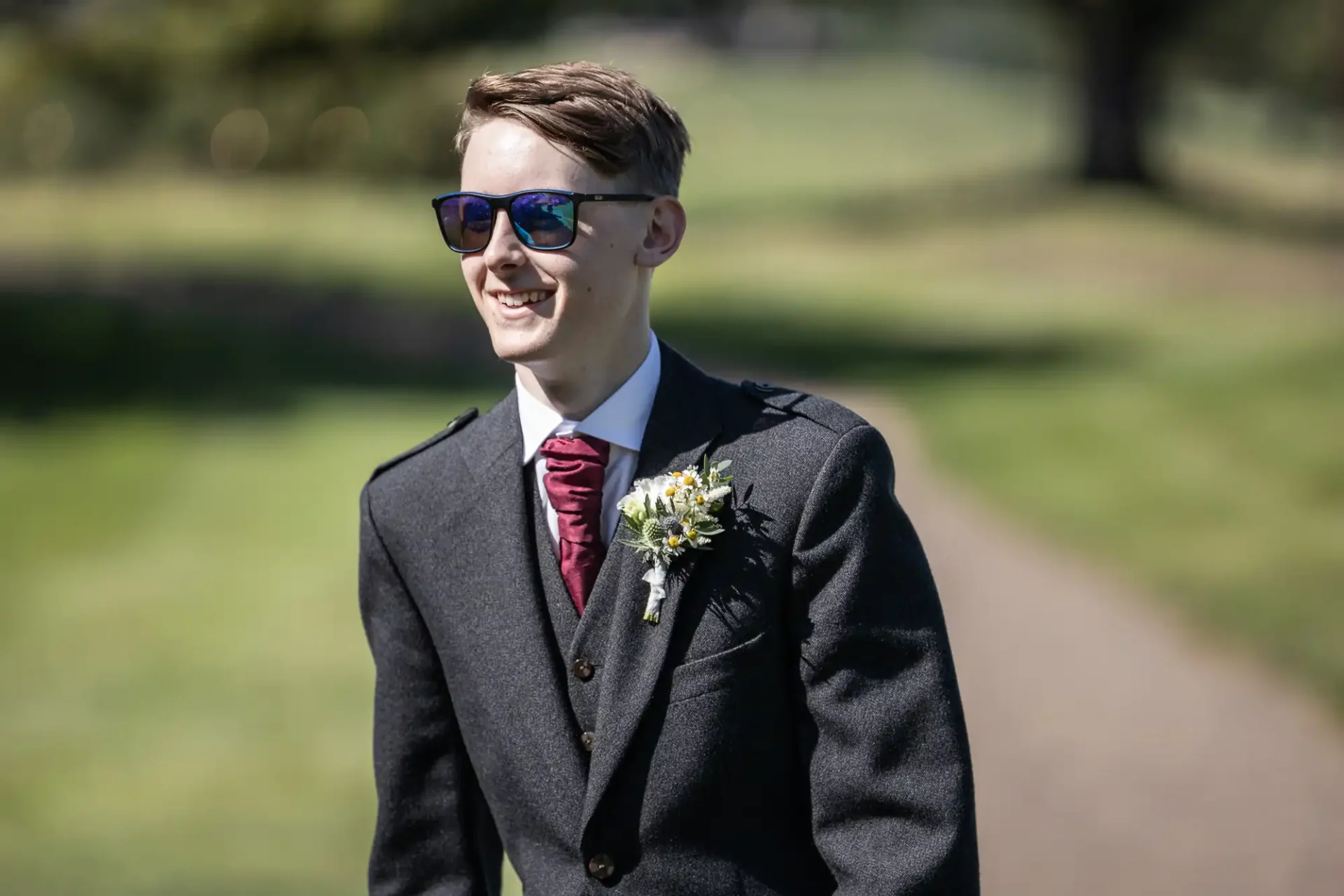 A man wearing sunglasses and a dark suit with a red tie and boutonniere stands outdoors on a sunny day, smiling.