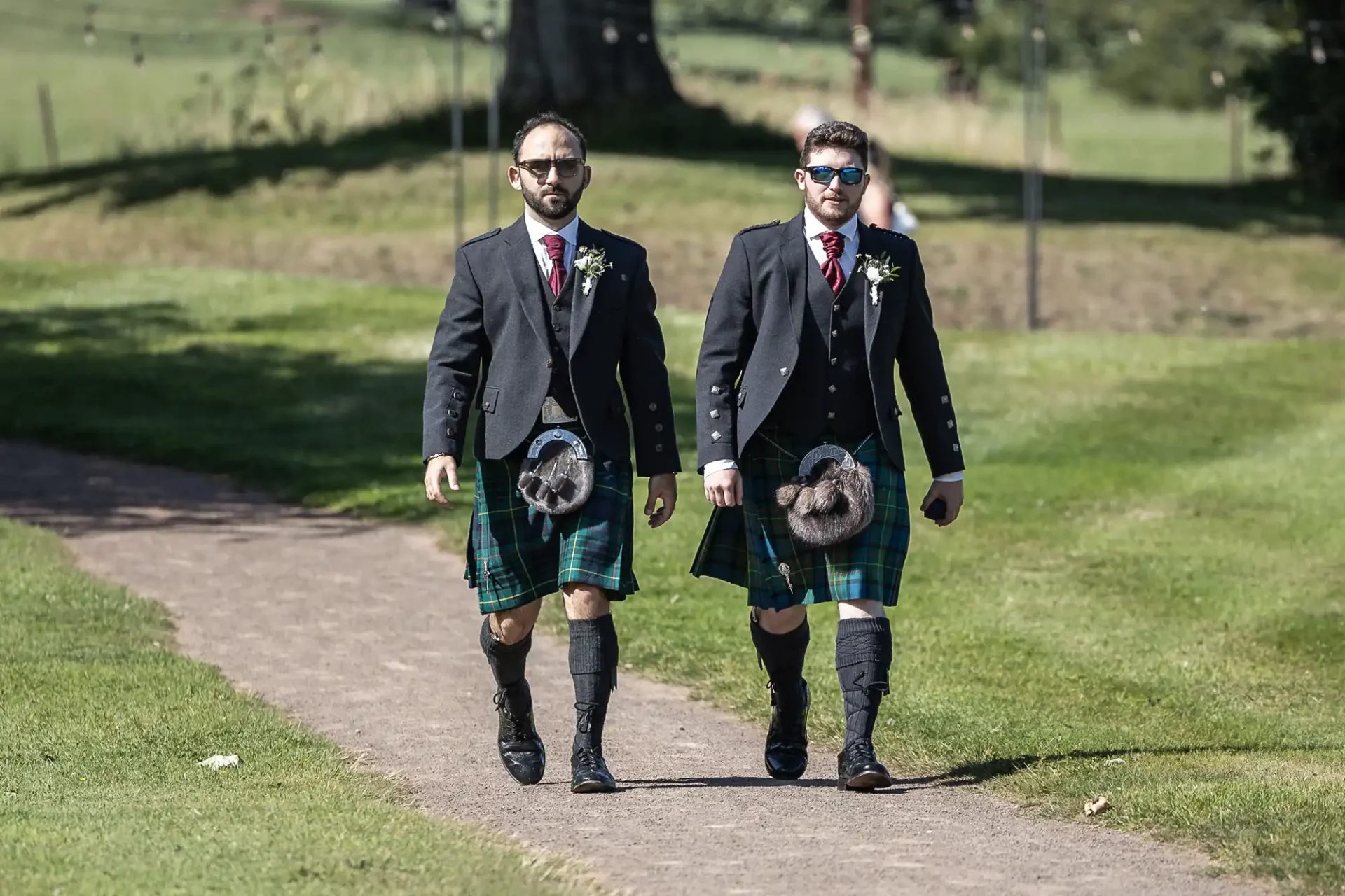 Two men in kilts and jackets walk on a path in a park, wearing sunglasses and floral boutonnières.