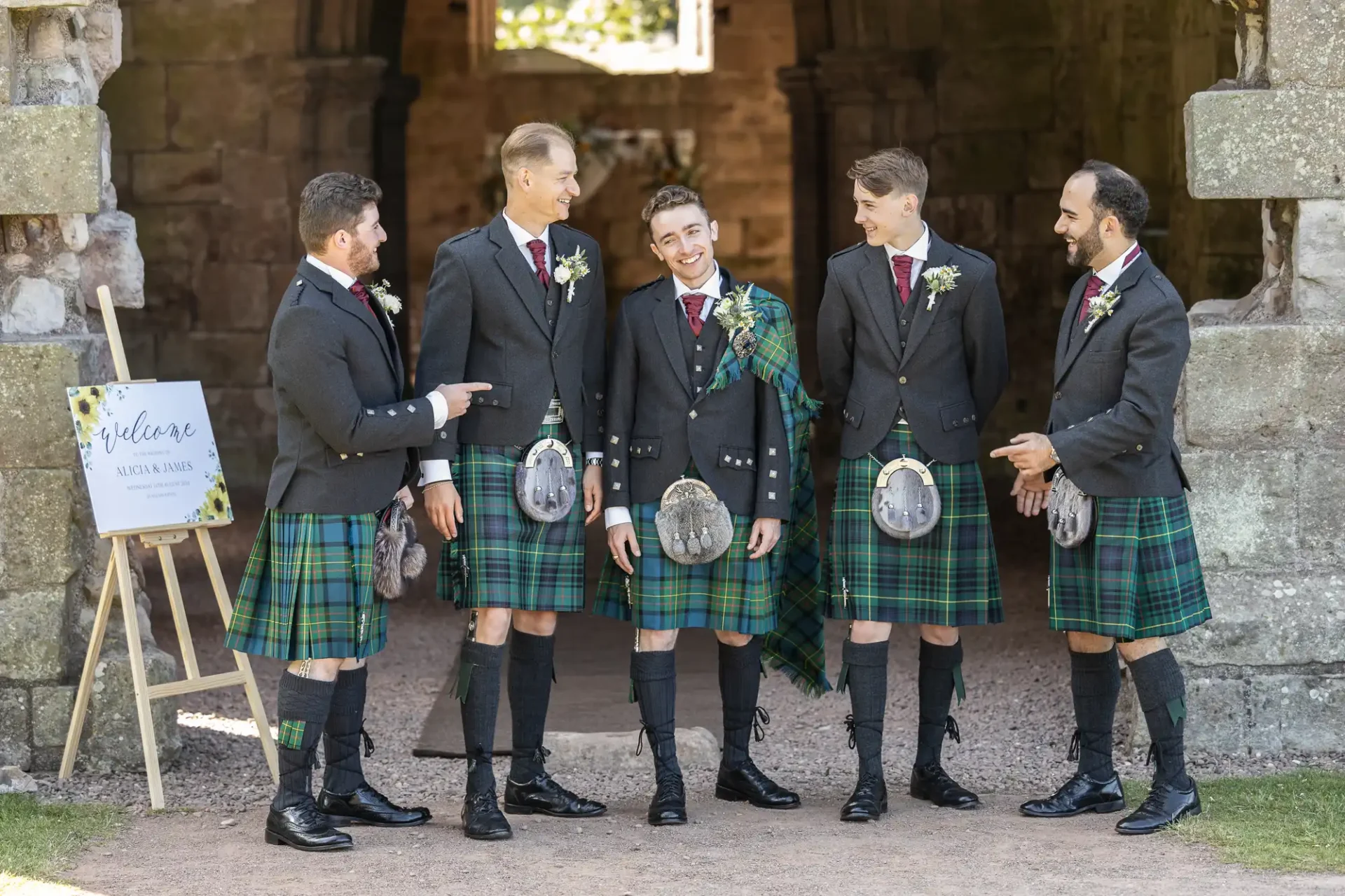 Five men in kilts and formal jackets stand together outside, smiling and talking. One holds a tartan sash, and there's a welcome sign nearby.