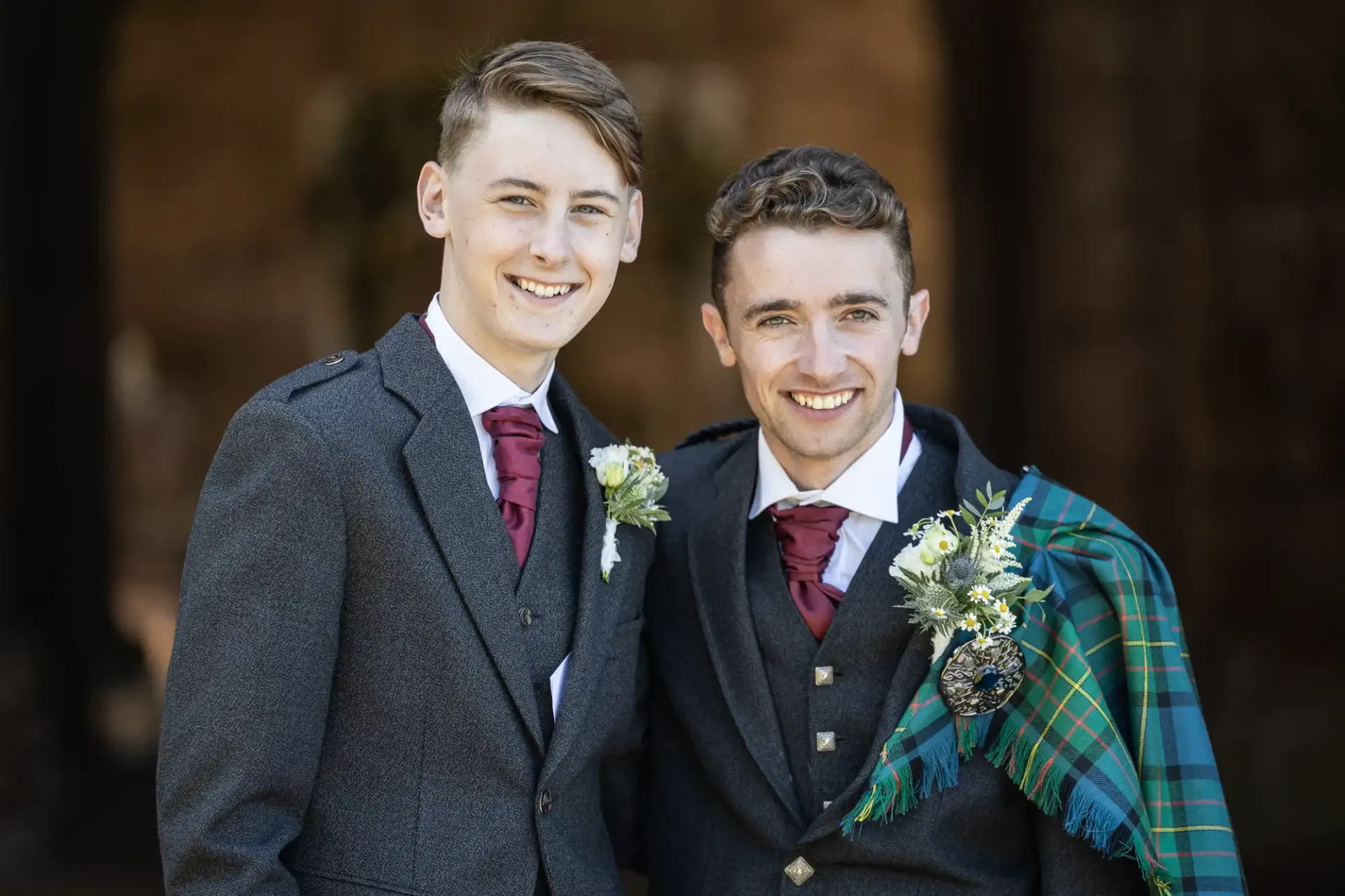 Two men in formal attire, one with a green tartan shawl, stand closely together outdoors. Both are smiling.