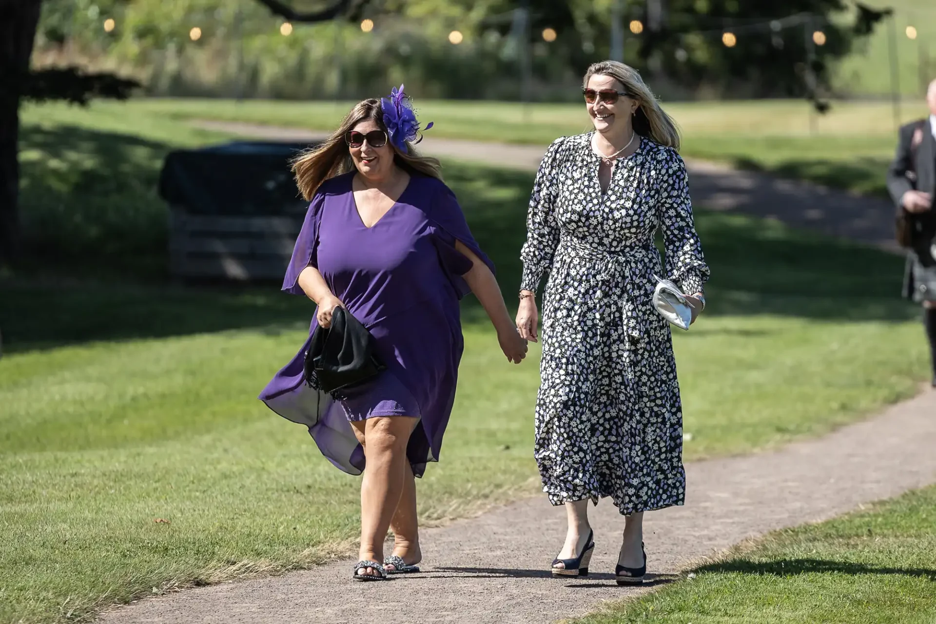 Two women walk hand in hand outside on a sunny day. One wears a purple dress with a fascinator, and the other a black and white patterned dress, holding a silver clutch.