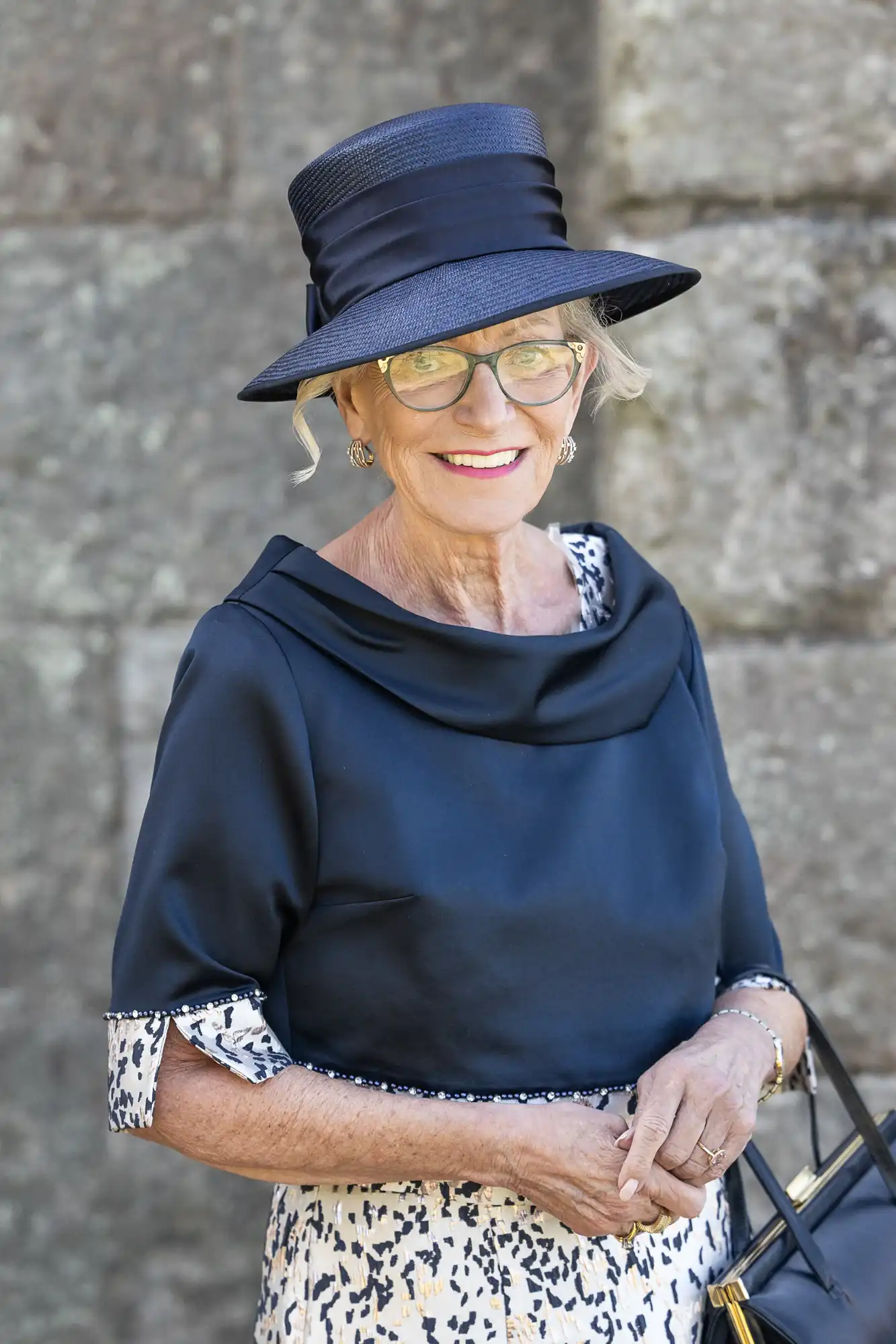 A woman wearing a black hat, glasses, and a navy outfit with a patterned skirt stands in front of a stone wall, smiling.