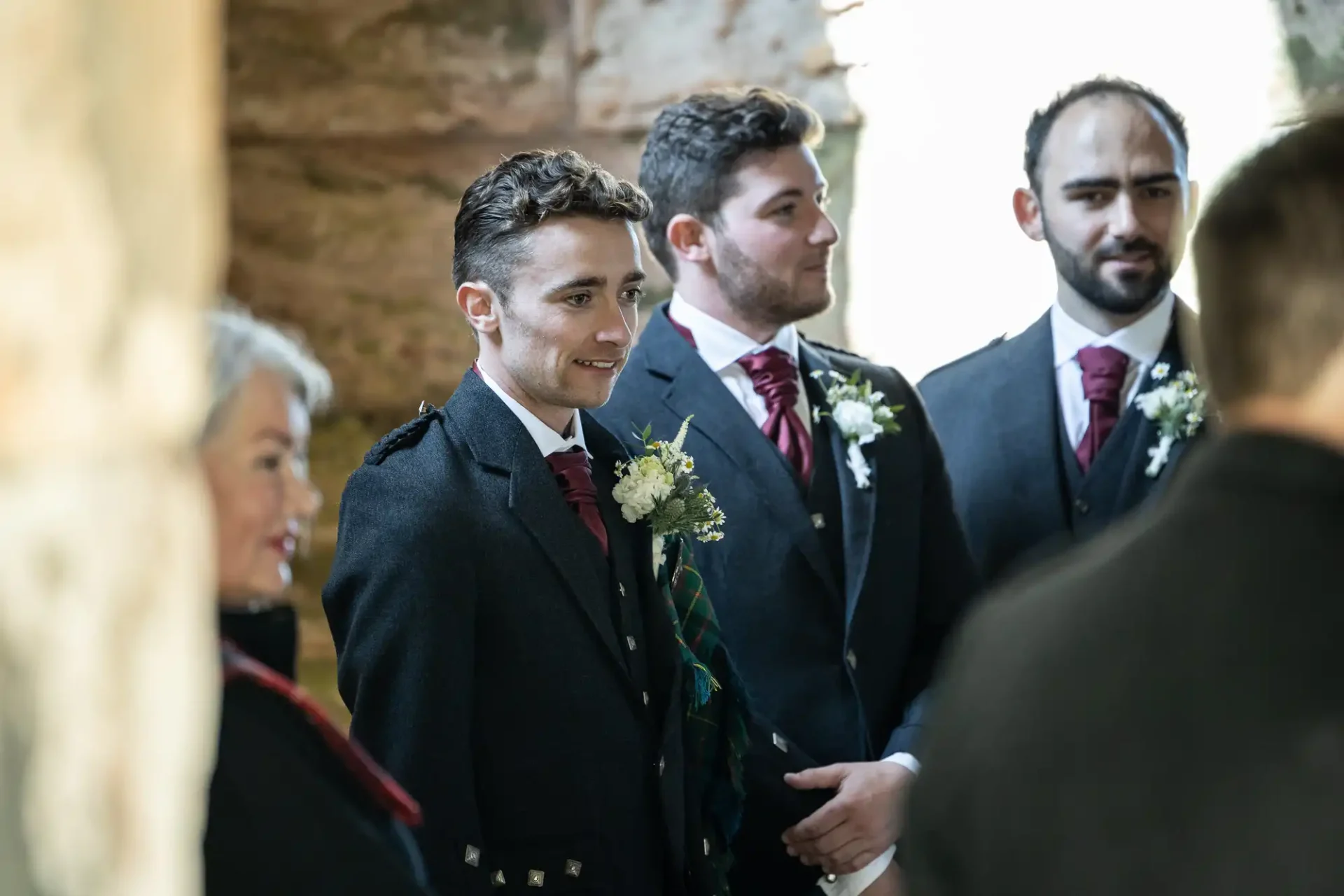 Three men in formal dark suits with red ties stand indoors. The central man wears a tartan sash. A woman to the left is slightly blurred. Stone walls are visible in the background.