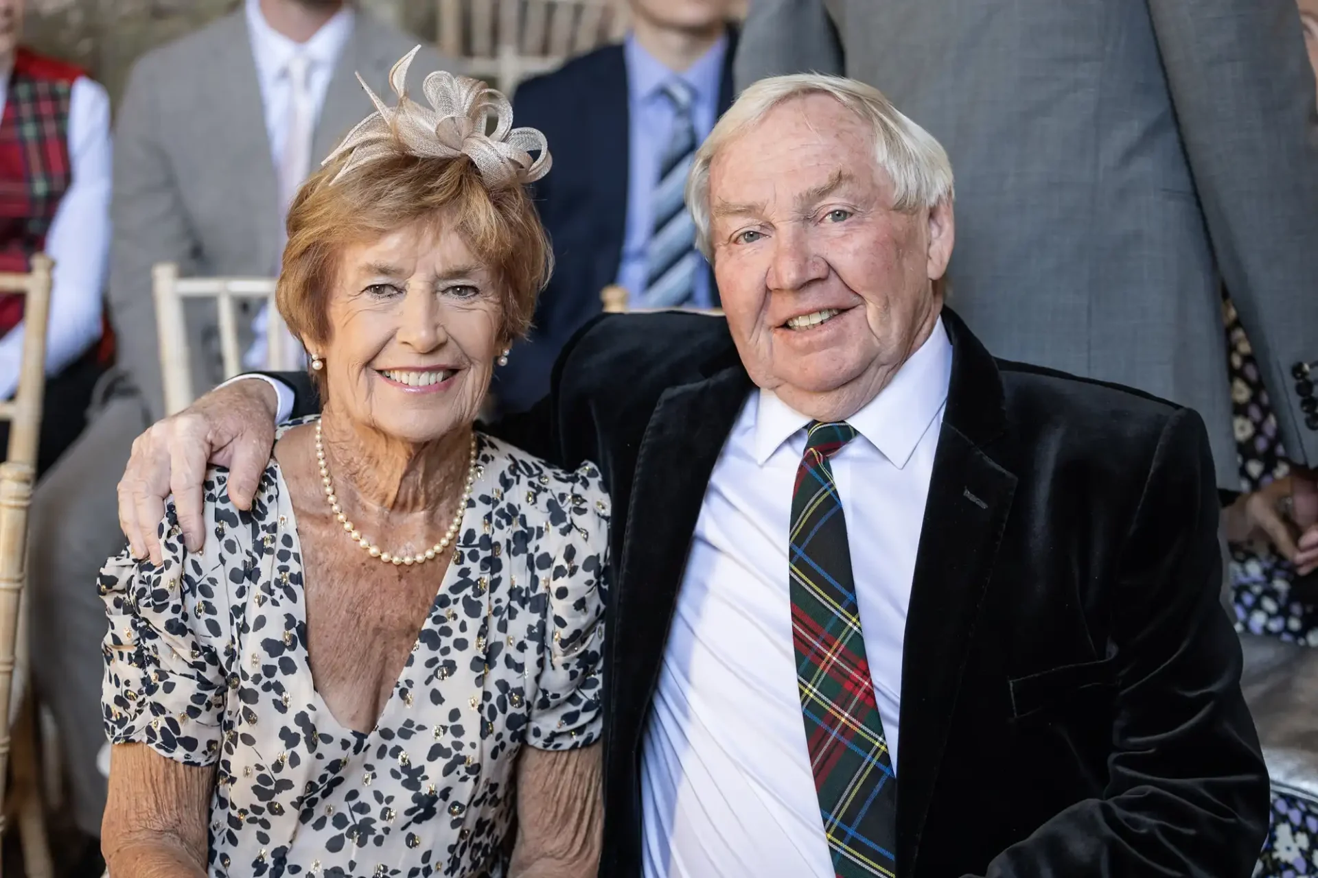 An elderly couple sitting together, smiling at an event. The woman is wearing a floral dress and fascinator, and the man is in a suit with a plaid tie.