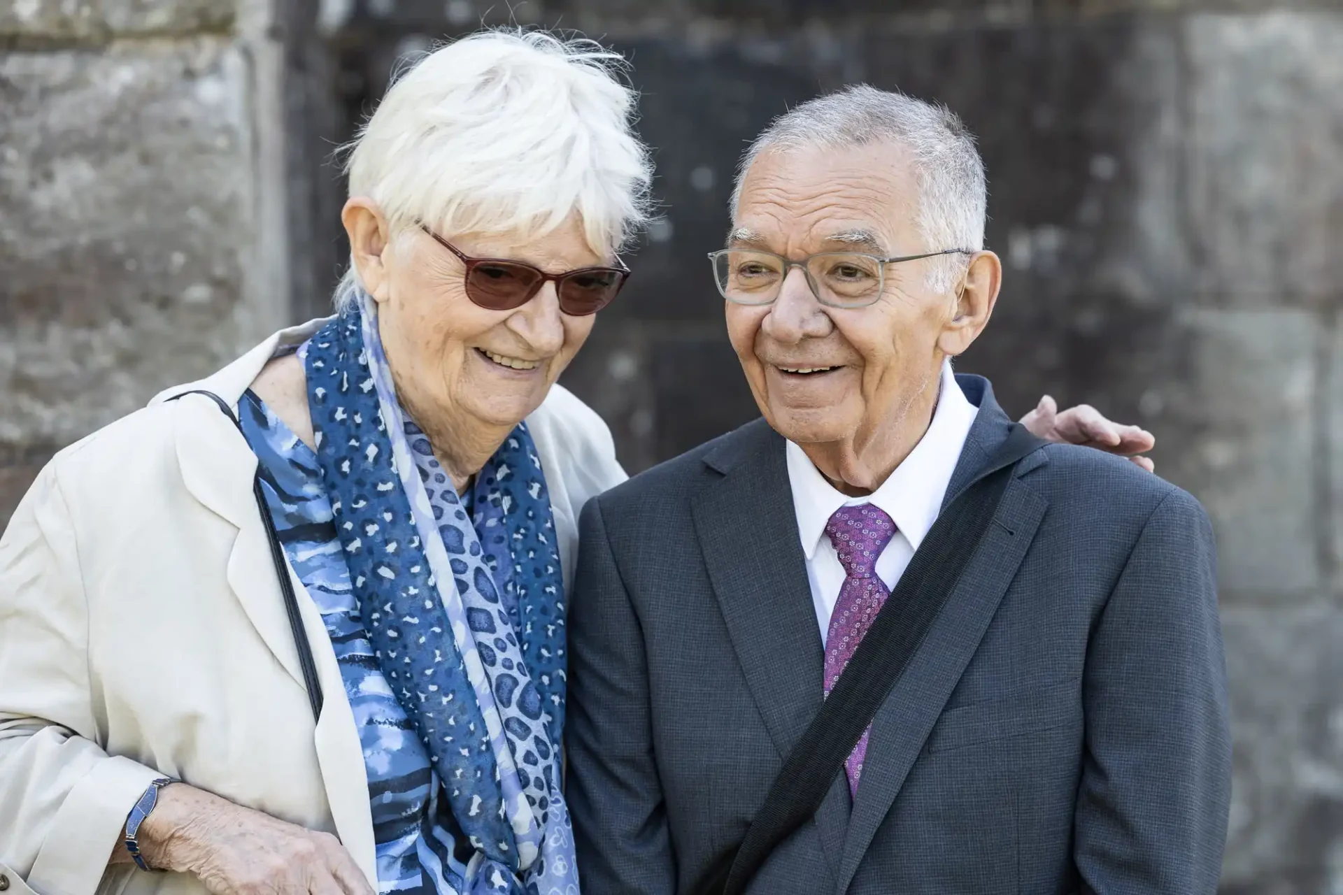 An elderly couple smiling, with the woman wearing glasses, a blue scarf, and a beige coat, and the man in a suit and purple tie, standing close together outside.