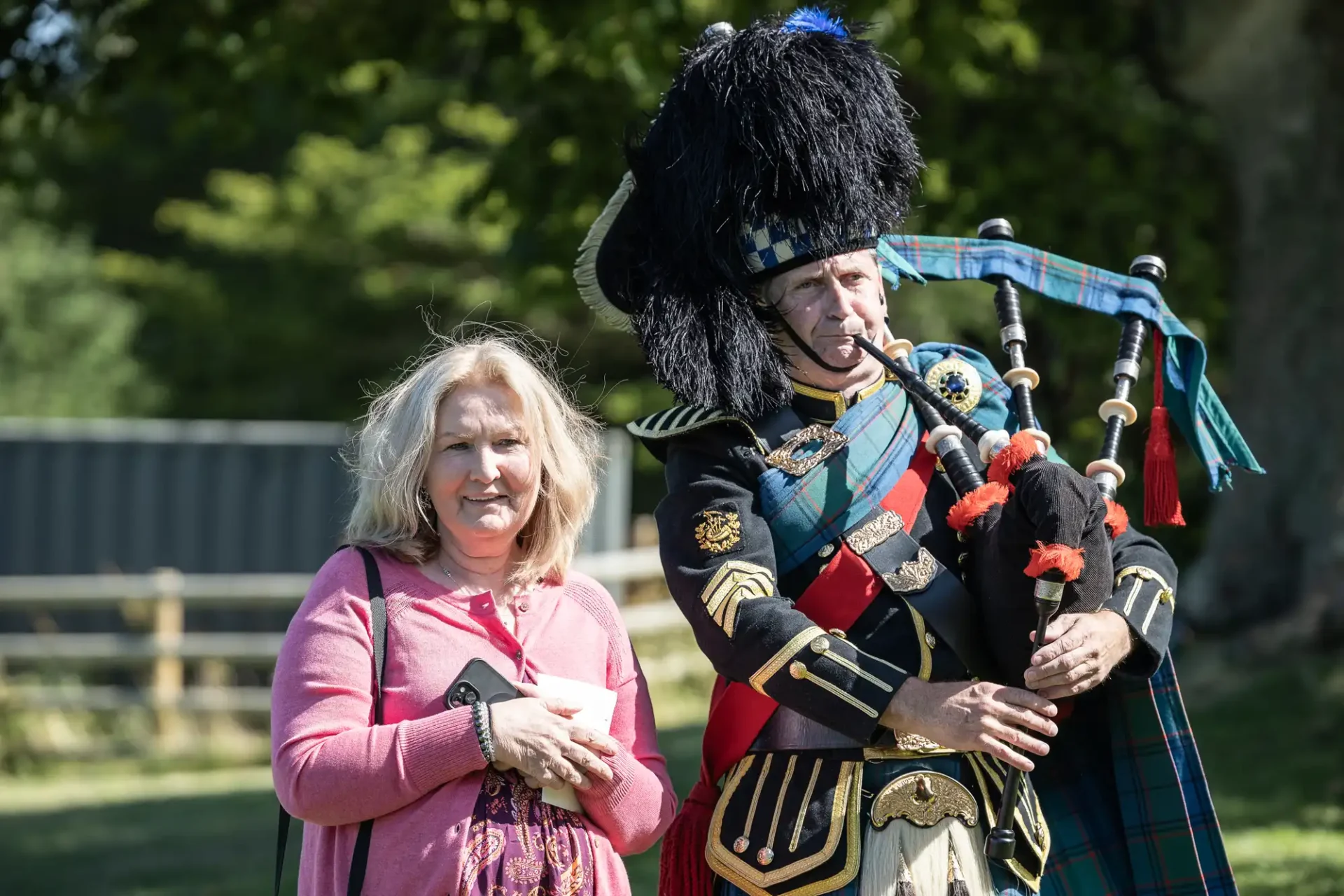 A woman stands beside a man in traditional Scottish attire playing bagpipes outdoors.