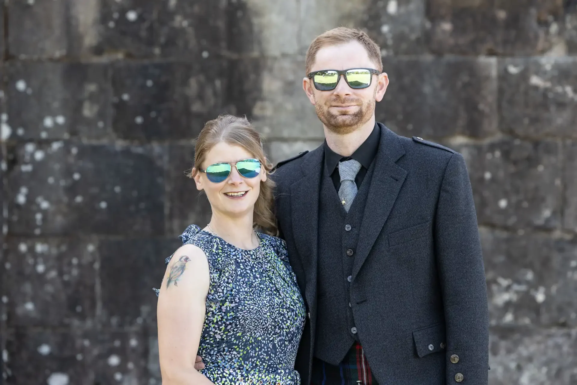 A couple wearing sunglasses stands in front of a stone wall. The man is in a dark suit, and the woman is in a patterned dress.