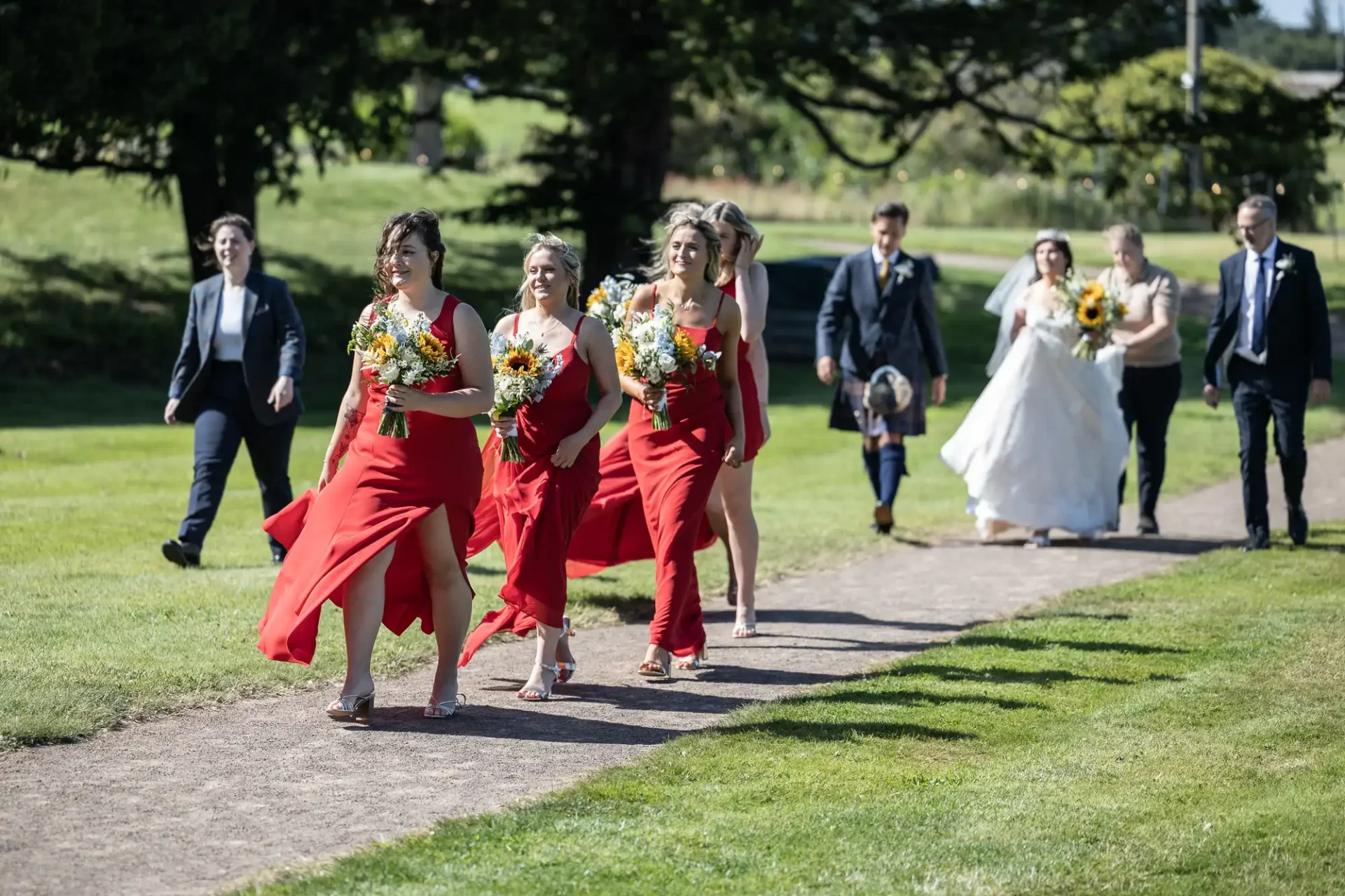 Bridal party walking on a park path. Bridesmaids in red dresses hold yellow flower bouquets; groomsmen and bride in the background.