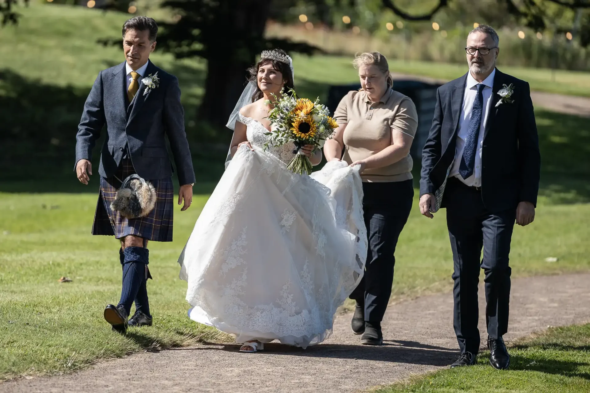 A bride in a white dress holds sunflowers, flanked by two men in suits and a woman in a beige shirt, walking on a path in a grassy area.