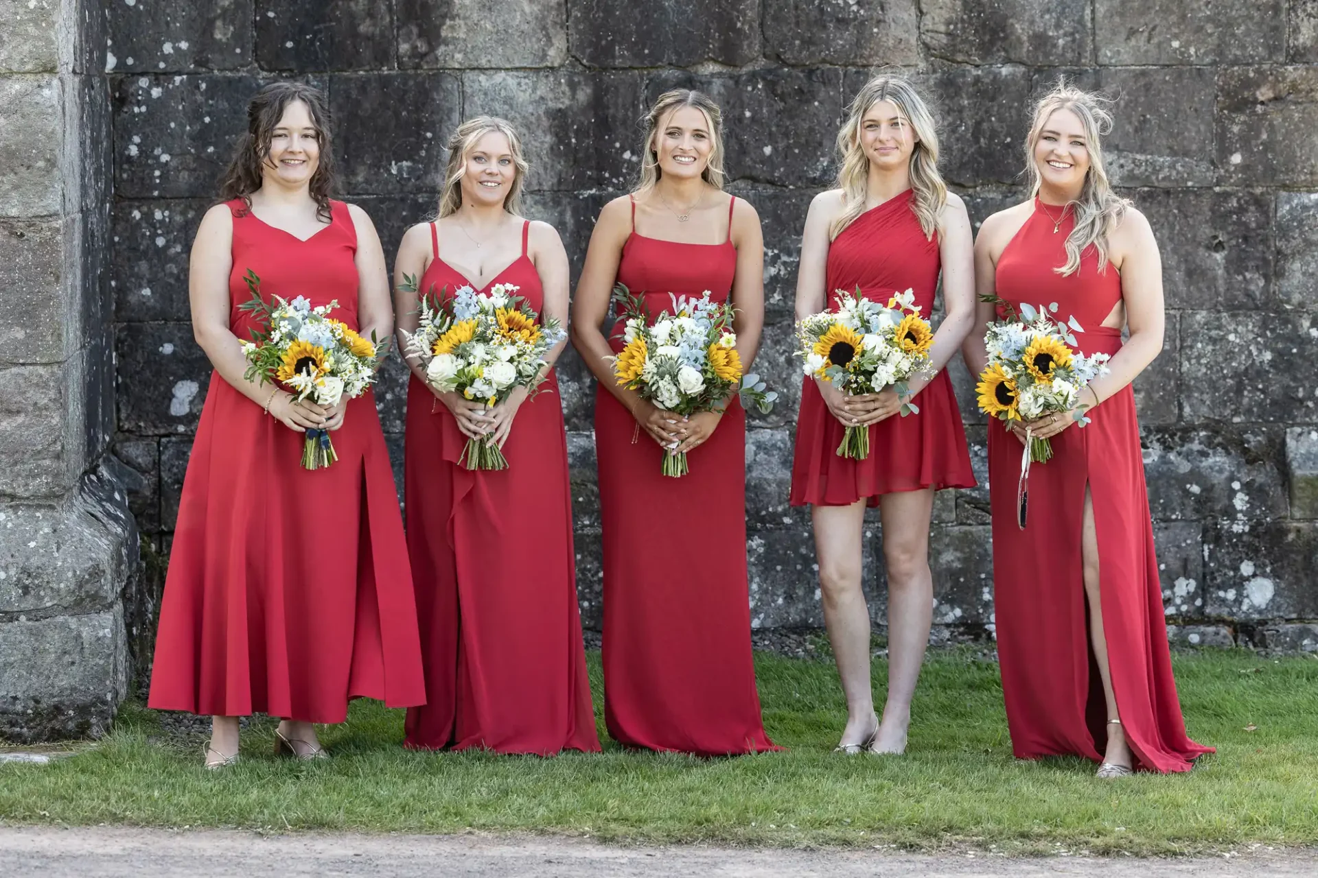 Five women in red dresses holding flower bouquets stand outside against a stone wall.