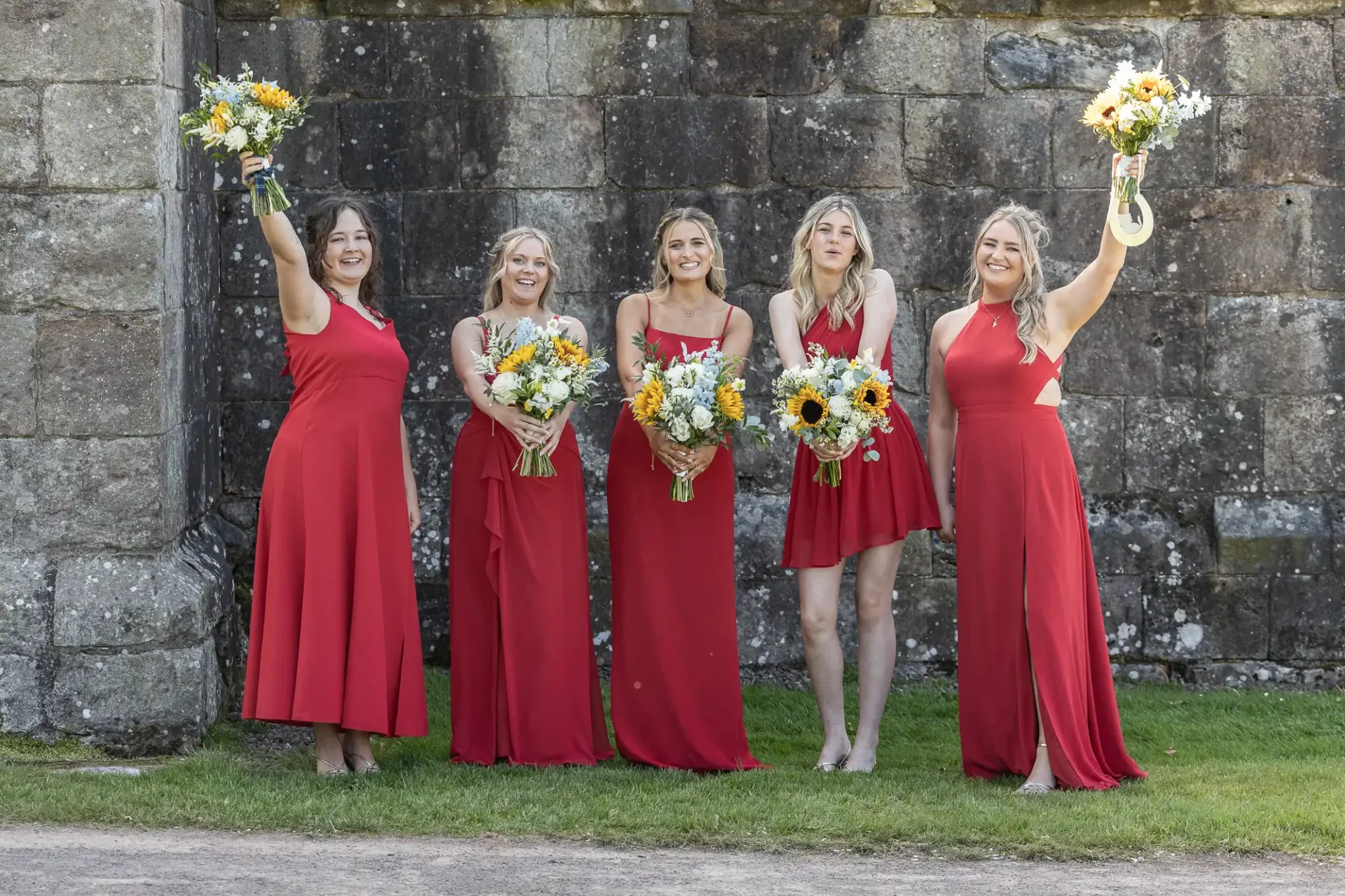 Five women in red dresses hold yellow flower bouquets, standing in front of a stone wall on grass.