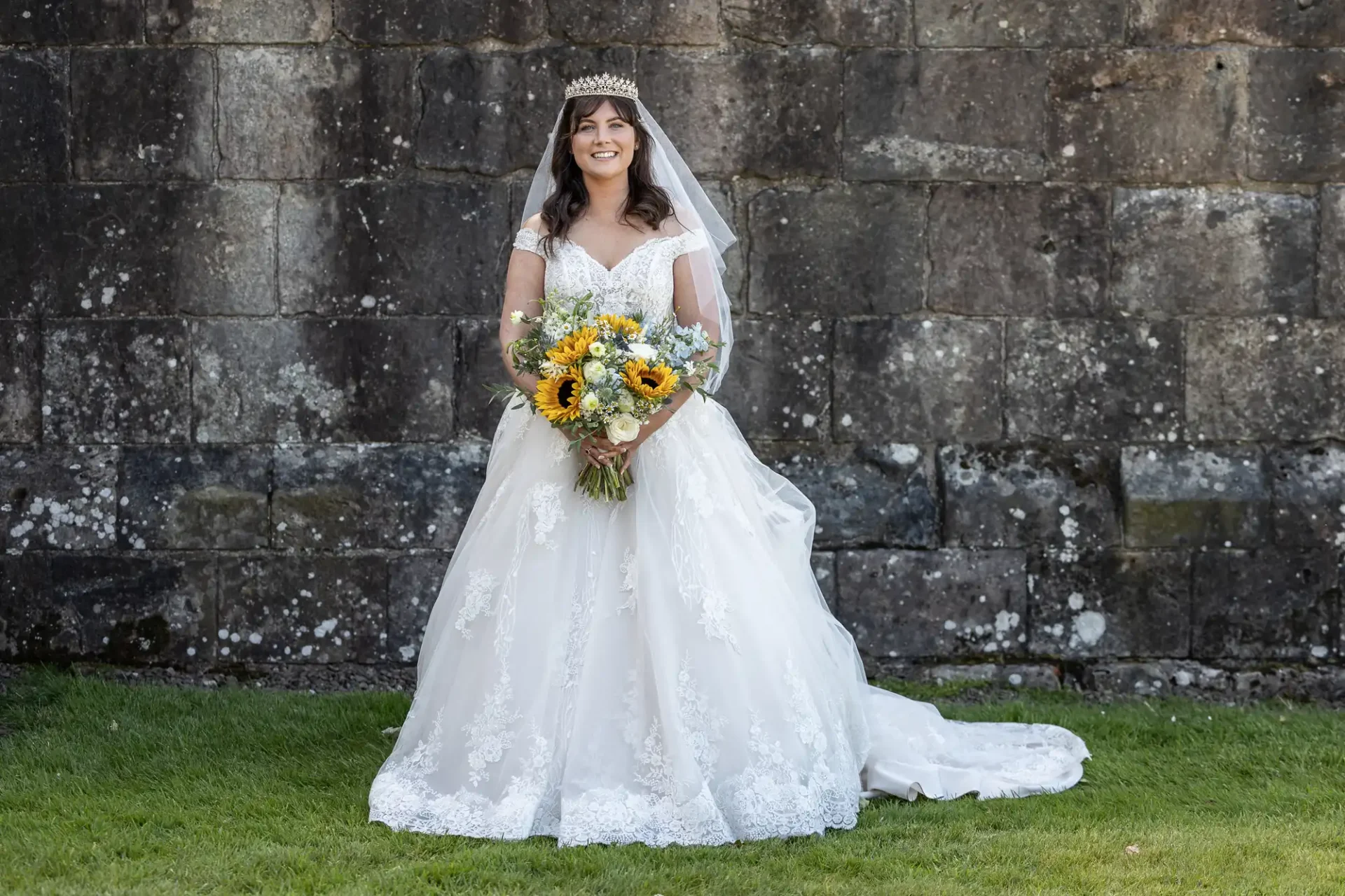 Bride in a white lace wedding dress holds a bouquet with sunflowers, standing on grass against a stone wall backdrop.