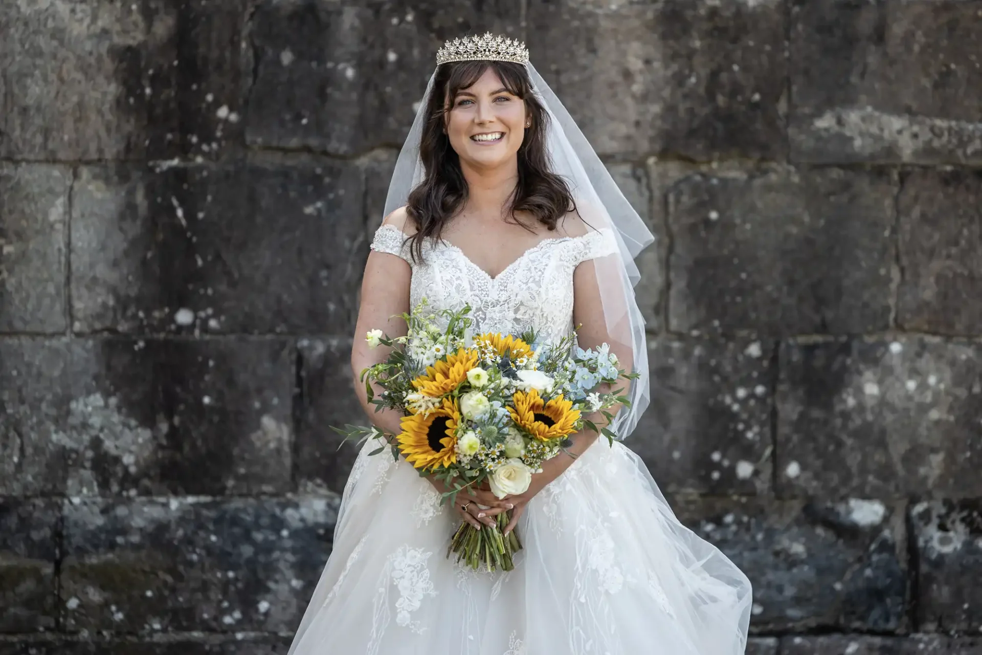 Bride smiling, wearing a white wedding dress with lace details and a tiara. She holds a bouquet of sunflowers in front of a stone wall.