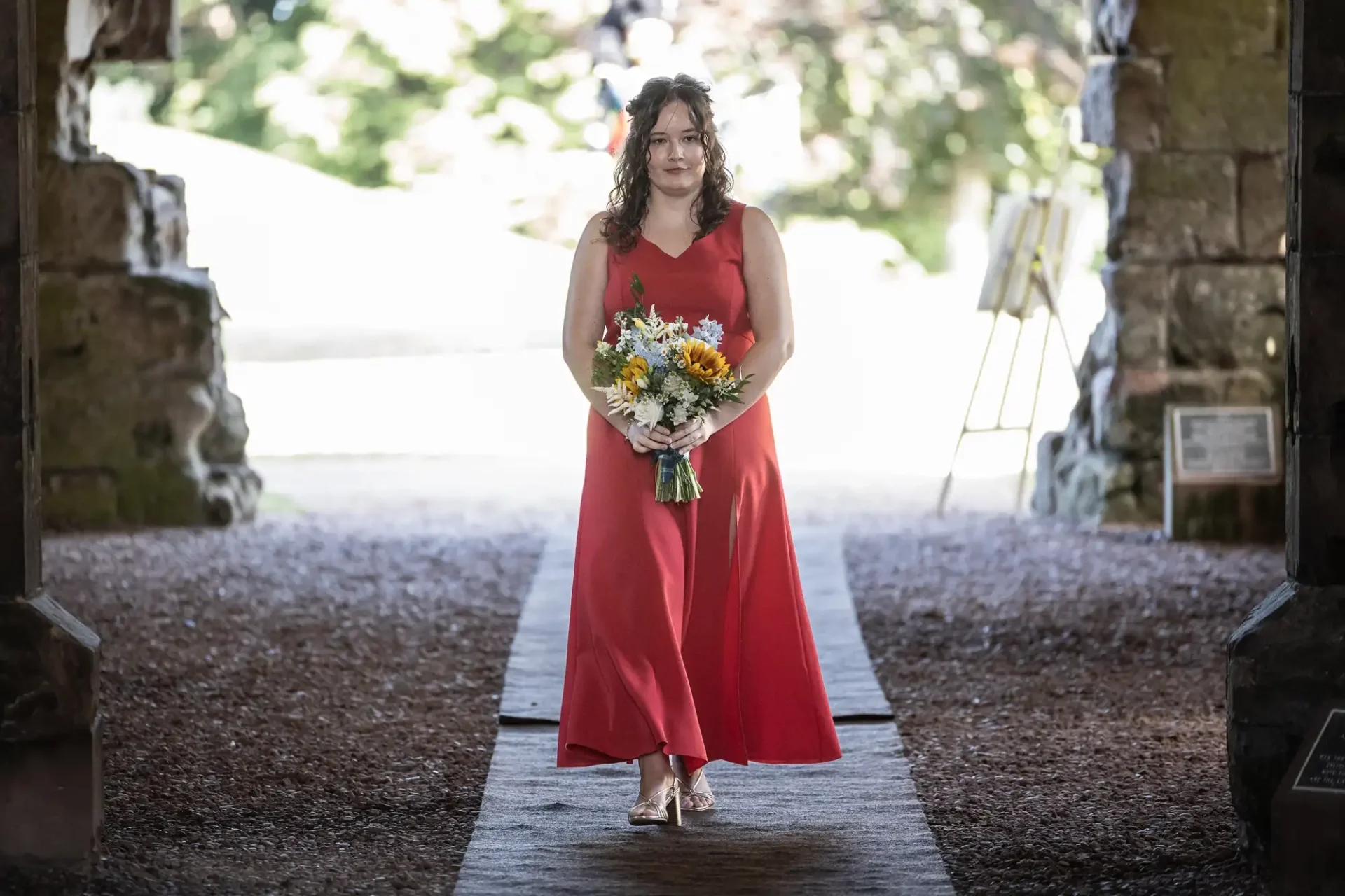 Woman in a red dress holding a bouquet walks down an outdoor aisle, surrounded by stone walls and greenery in the background.