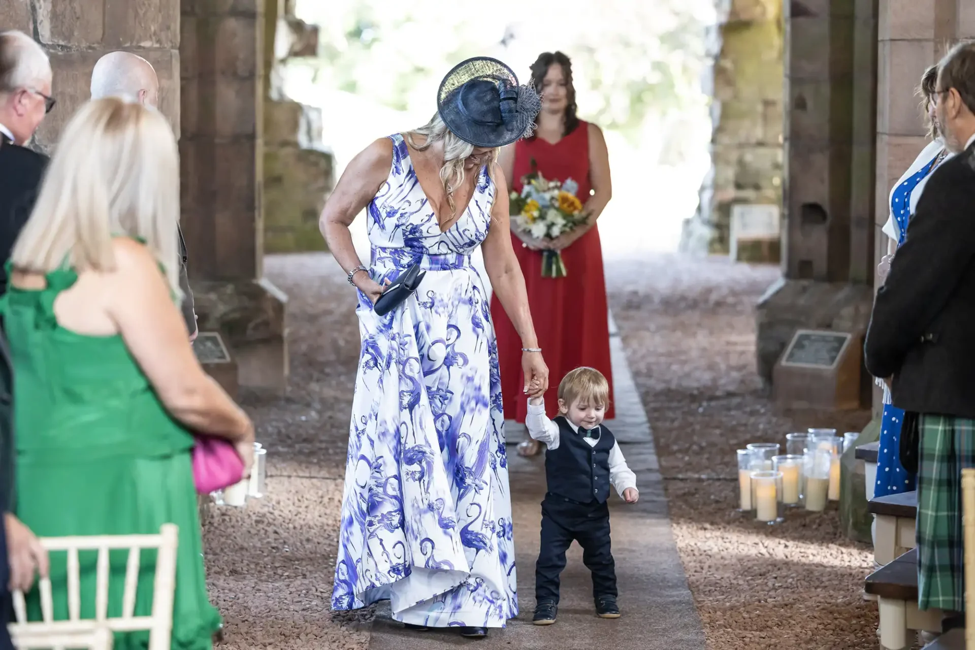 A woman in a floral dress walks down an aisle with a toddler in a suit at a wedding ceremony.