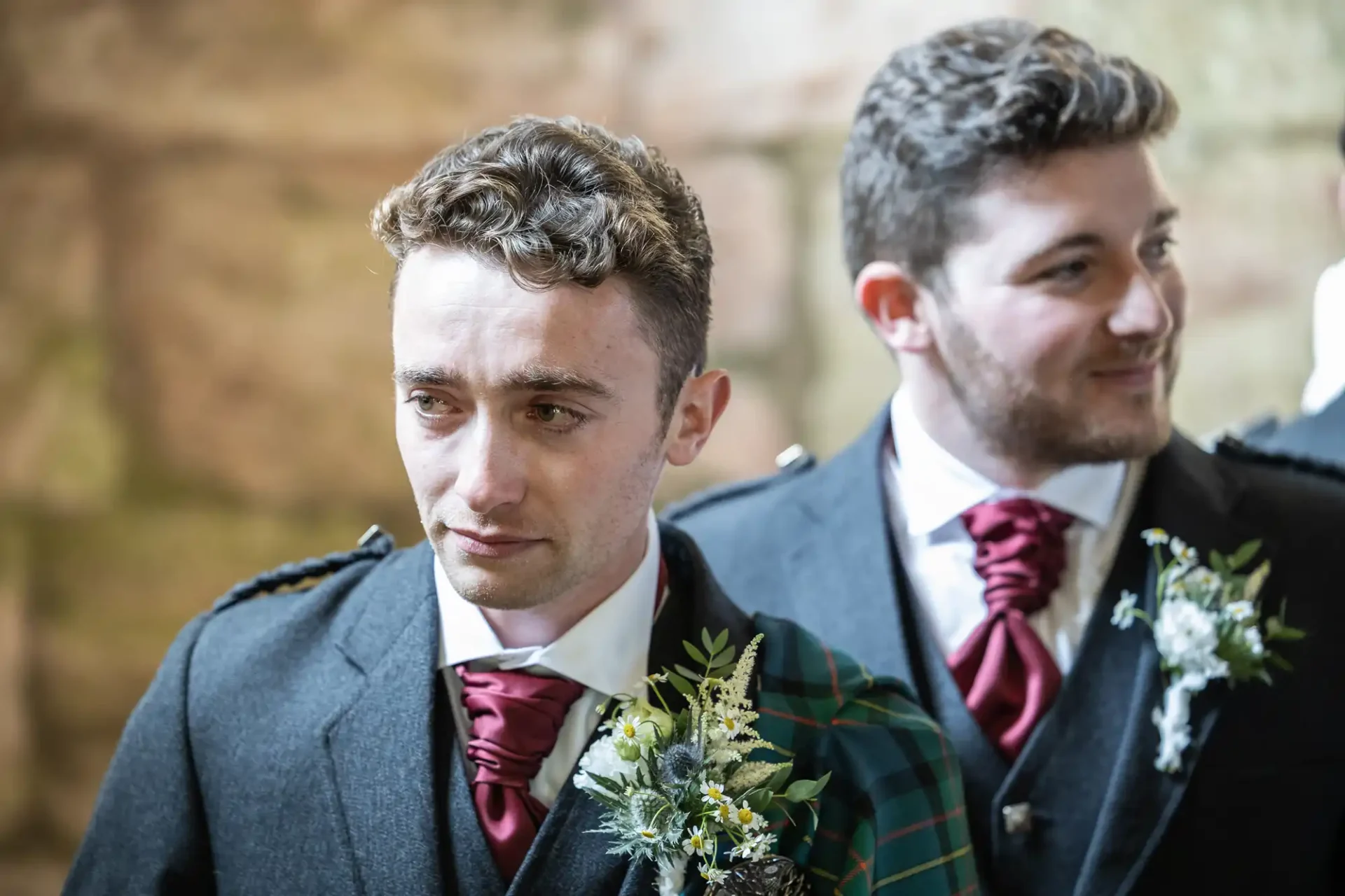 Two men in formal attire, with maroon ties and floral lapel decorations, stand side by side indoors. One wears a tartan sash. Background is blurred.