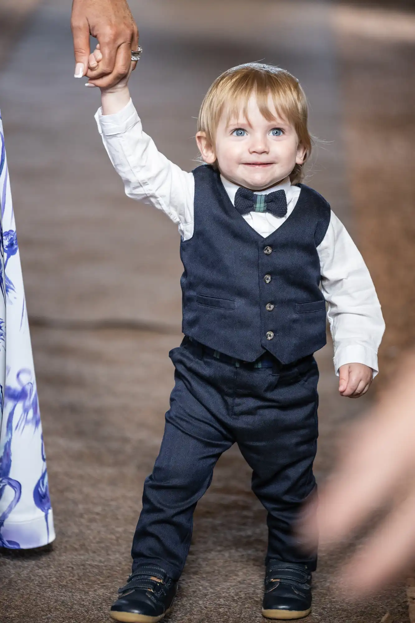 A young child in formal attire, including a bow tie and vest, walks holding an adult's hand on a runway.