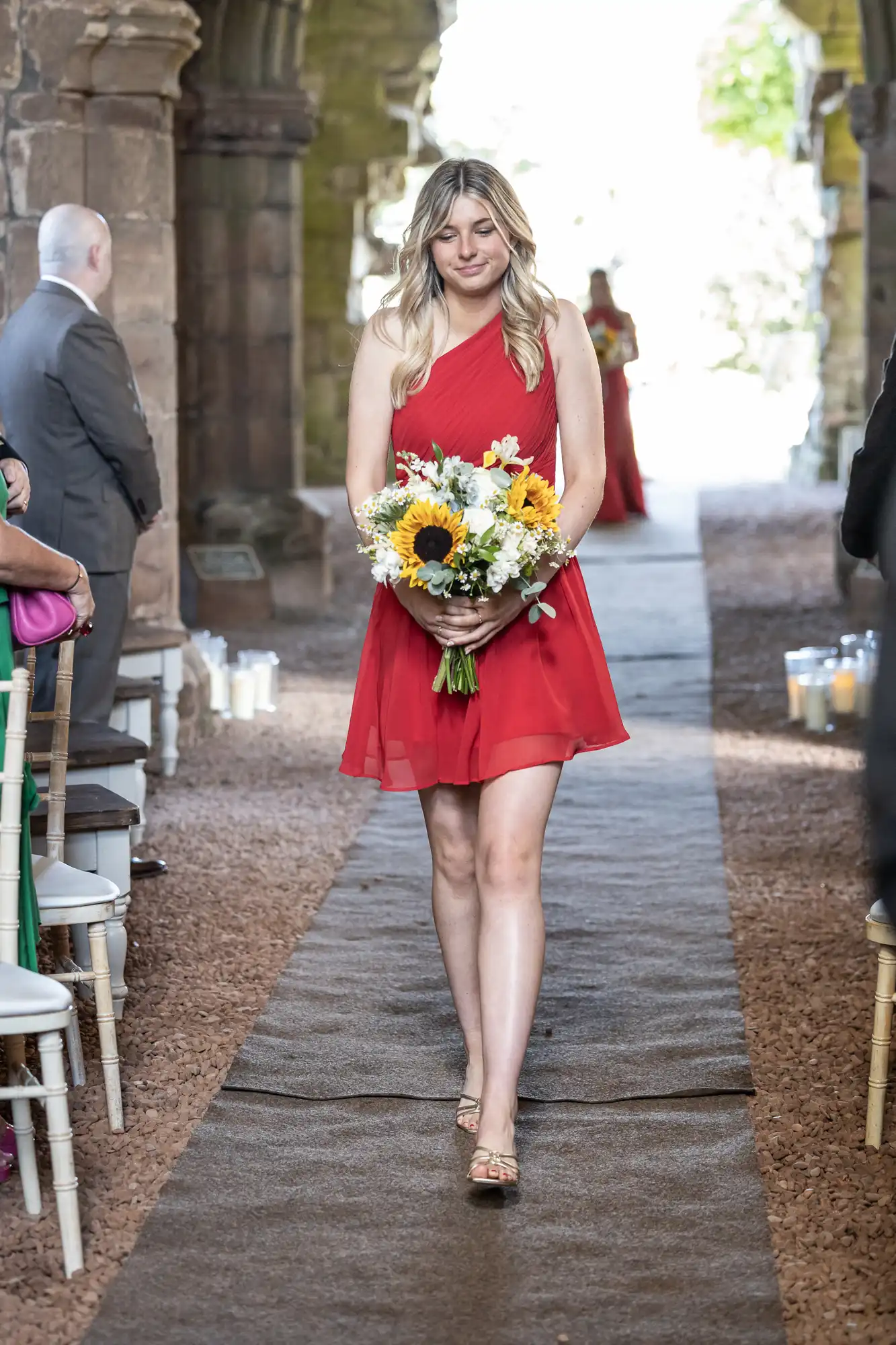 A woman in a red dress walks down an outdoor aisle holding a bouquet of sunflowers. Other people are seated nearby.