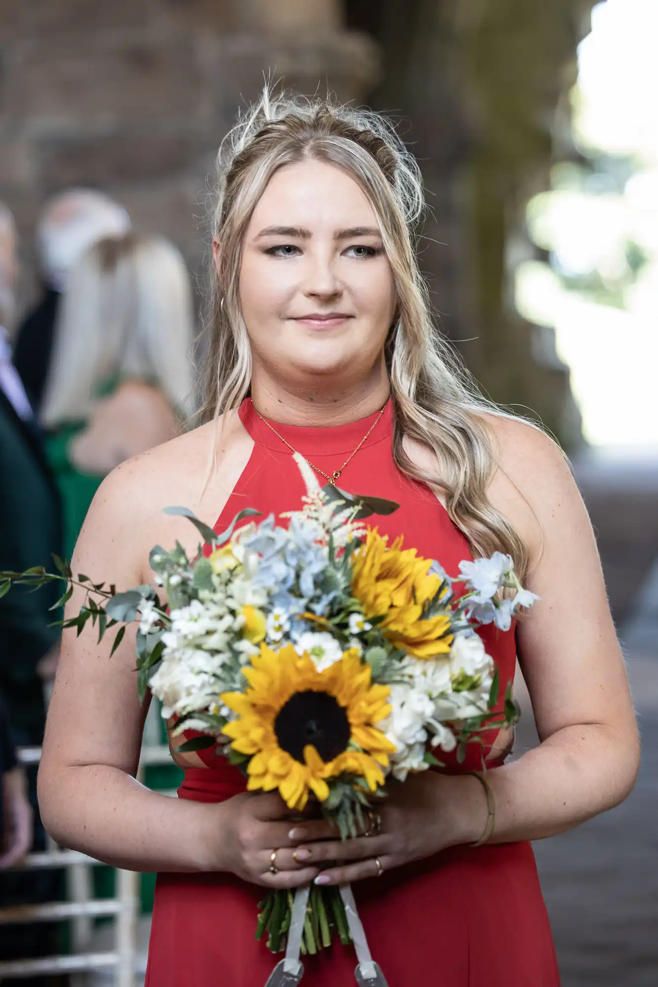 A woman in a red dress holds a bouquet with sunflowers and white flowers, standing outdoors.