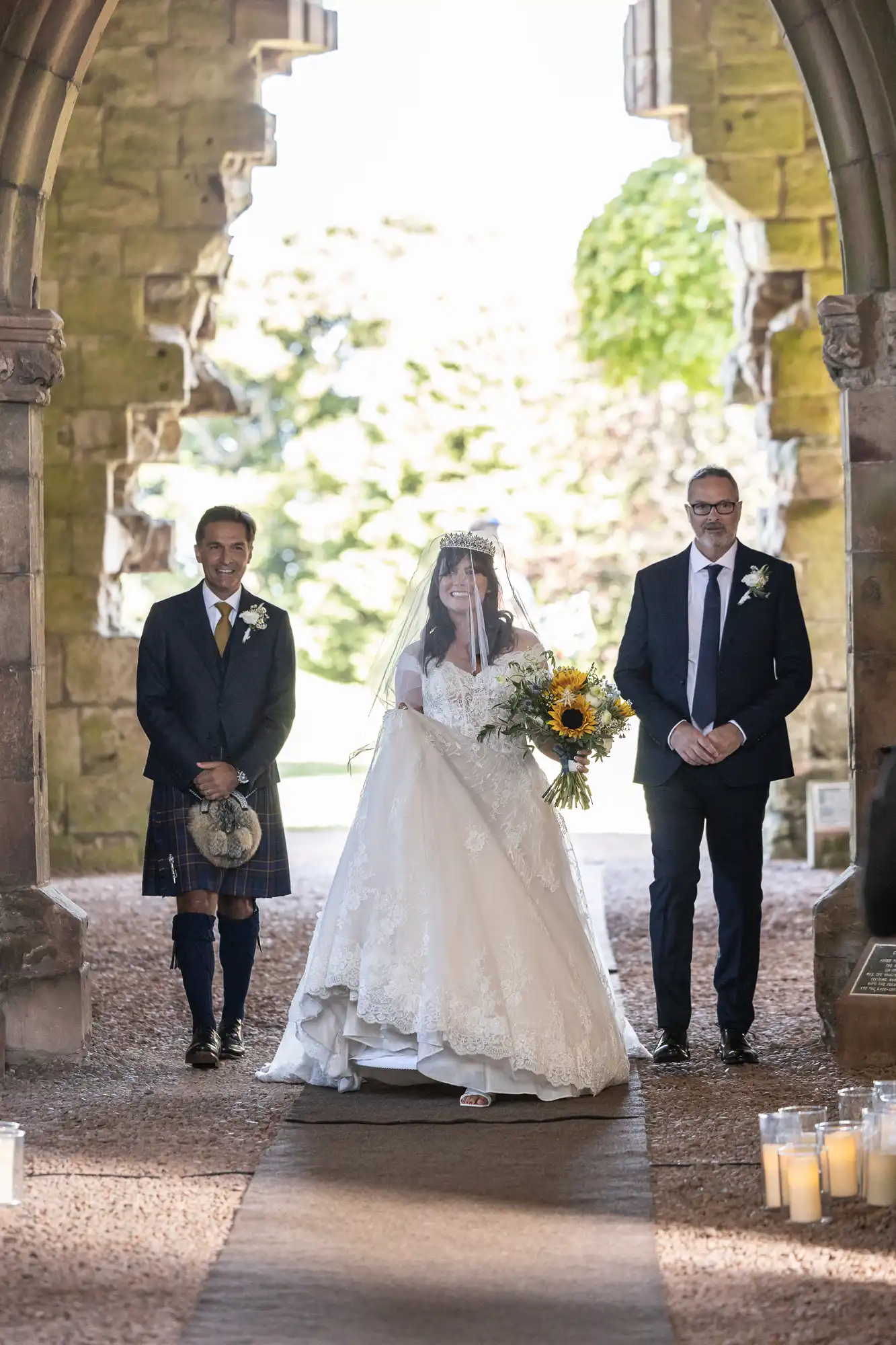 Bride in a white gown with veil holding a sunflower bouquet, walking down the aisle with two men in suits, under a stone arch, with candles on the floor.