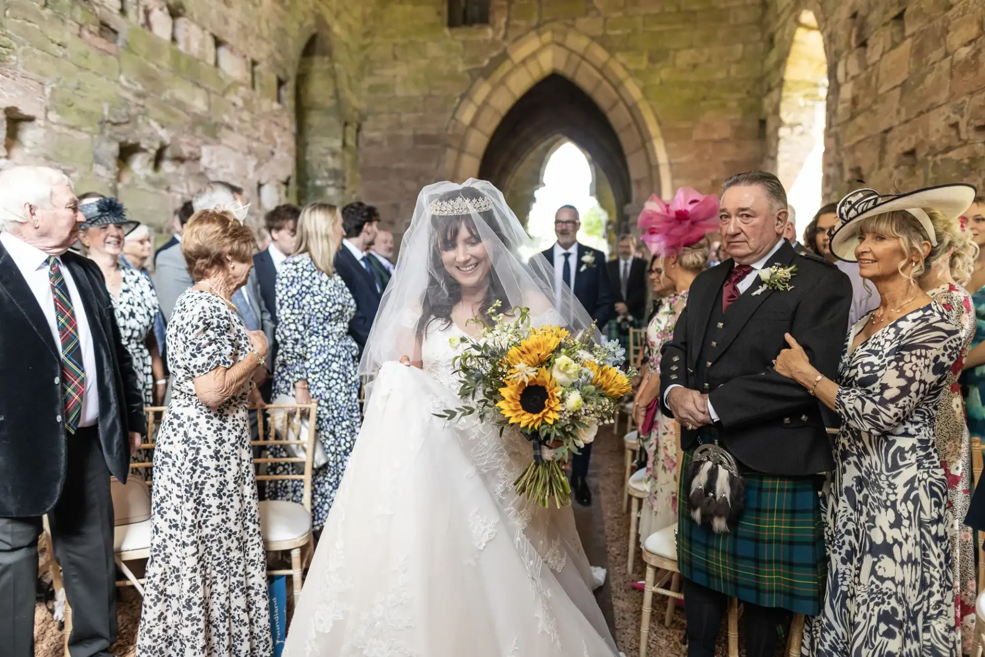 A bride in a white gown and veil holds sunflowers while walking down the aisle of an old stone building, accompanied by an older man. Guests stand on either side, some wearing hats.