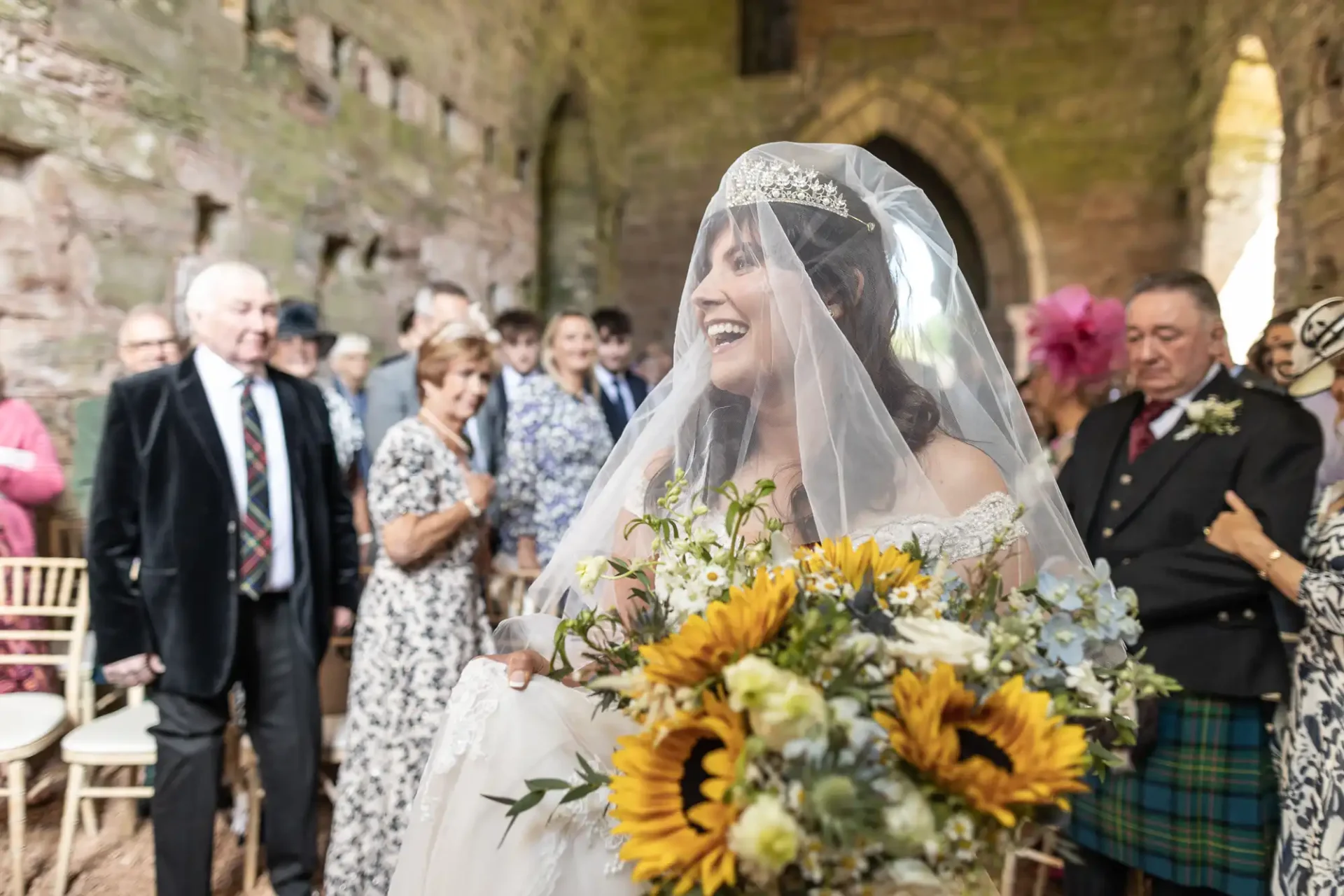 A bride in a veil and tiara holds a bouquet of sunflowers, walking down an aisle in a rustic stone venue. Guests, including a man in a kilt, stand and watch.