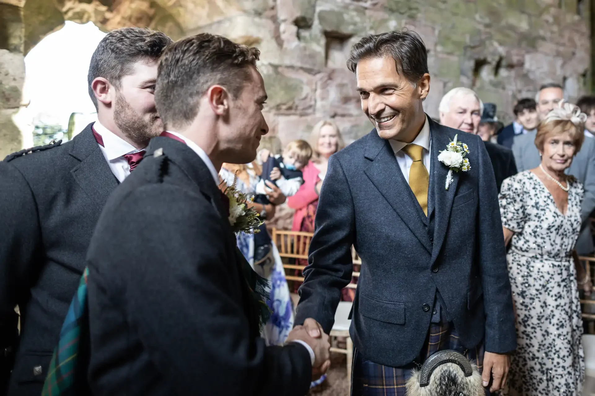 Two men in suits shake hands at a social gathering in a rustic stone setting, surrounded by seated guests.