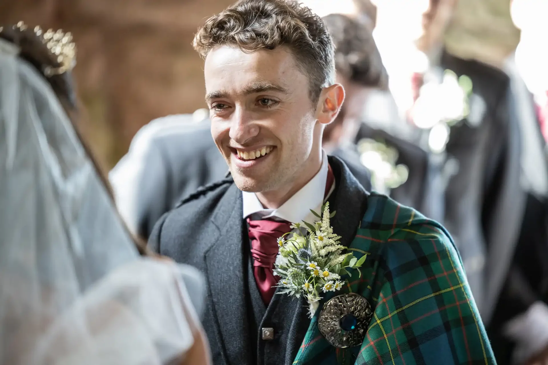 A man in a suit with a tartan sash smiling at a wedding ceremony.