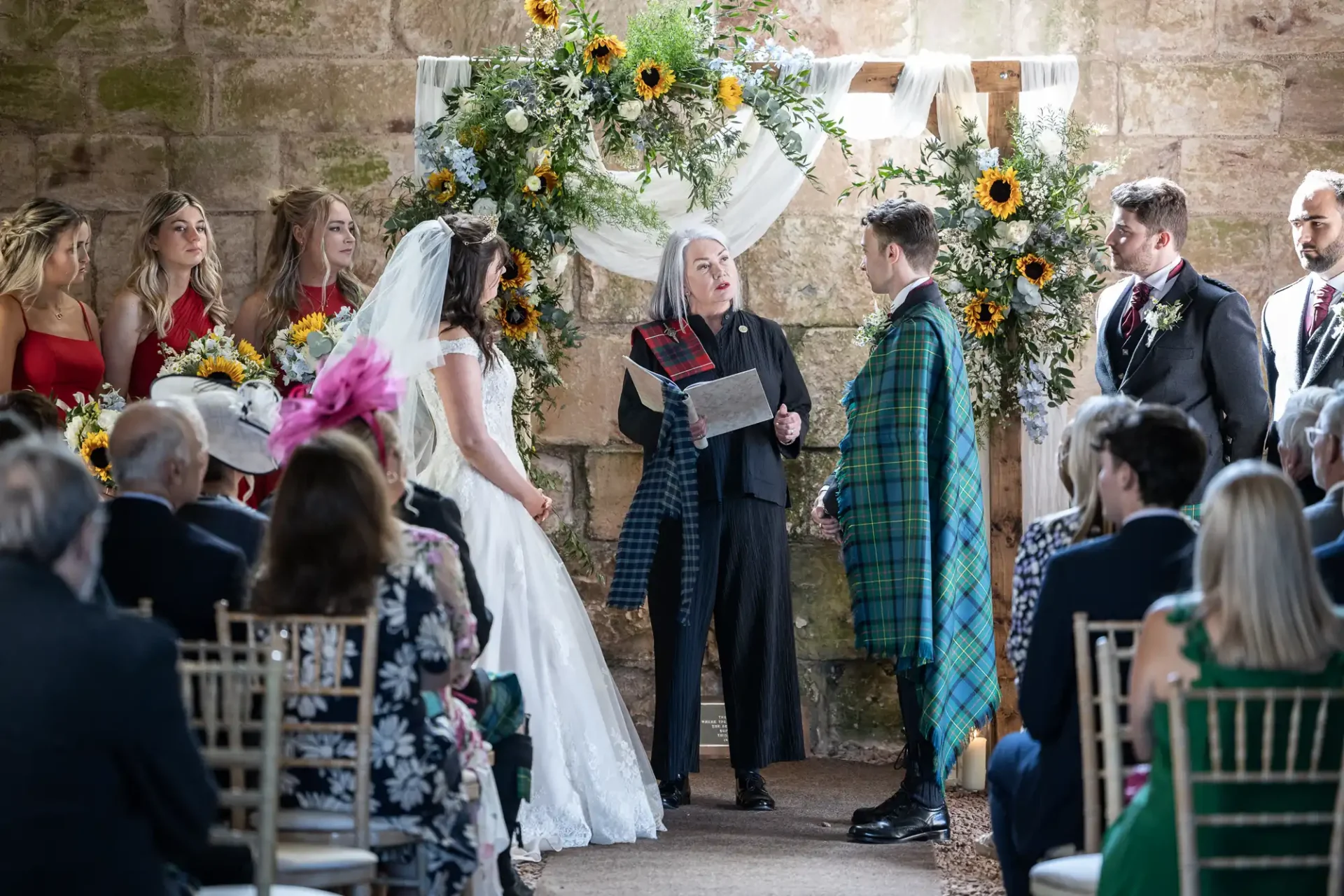 A couple stands before an officiant at a wedding ceremony. The bride is in a white dress, and the groom wears a suit with a tartan sash. Bridesmaids are in red dresses holding yellow flowers.