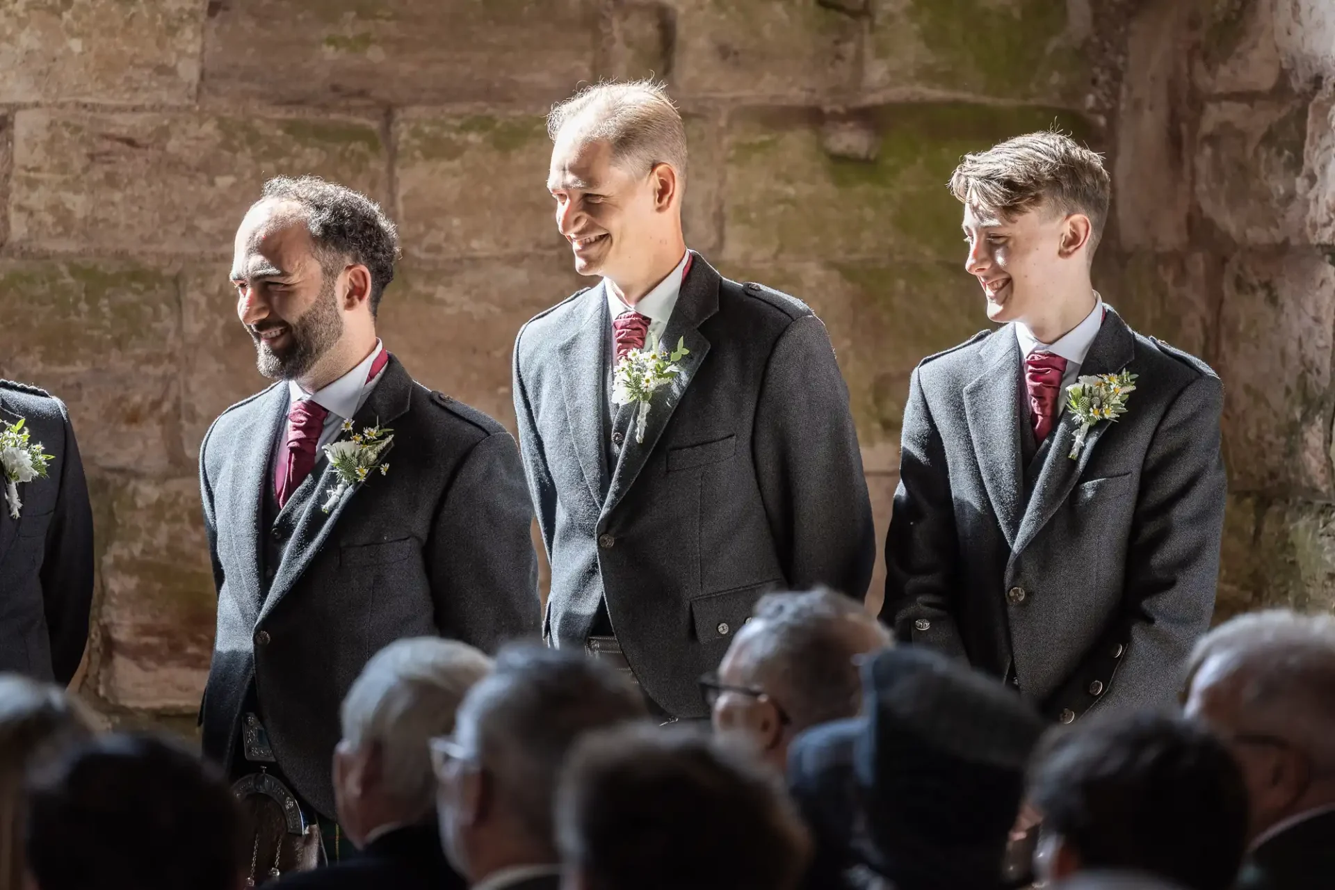 Three men in formal attire with matching grey suits and floral boutonnieres stand inside a rustic setting. They appear to be at an event with seated guests in the foreground.