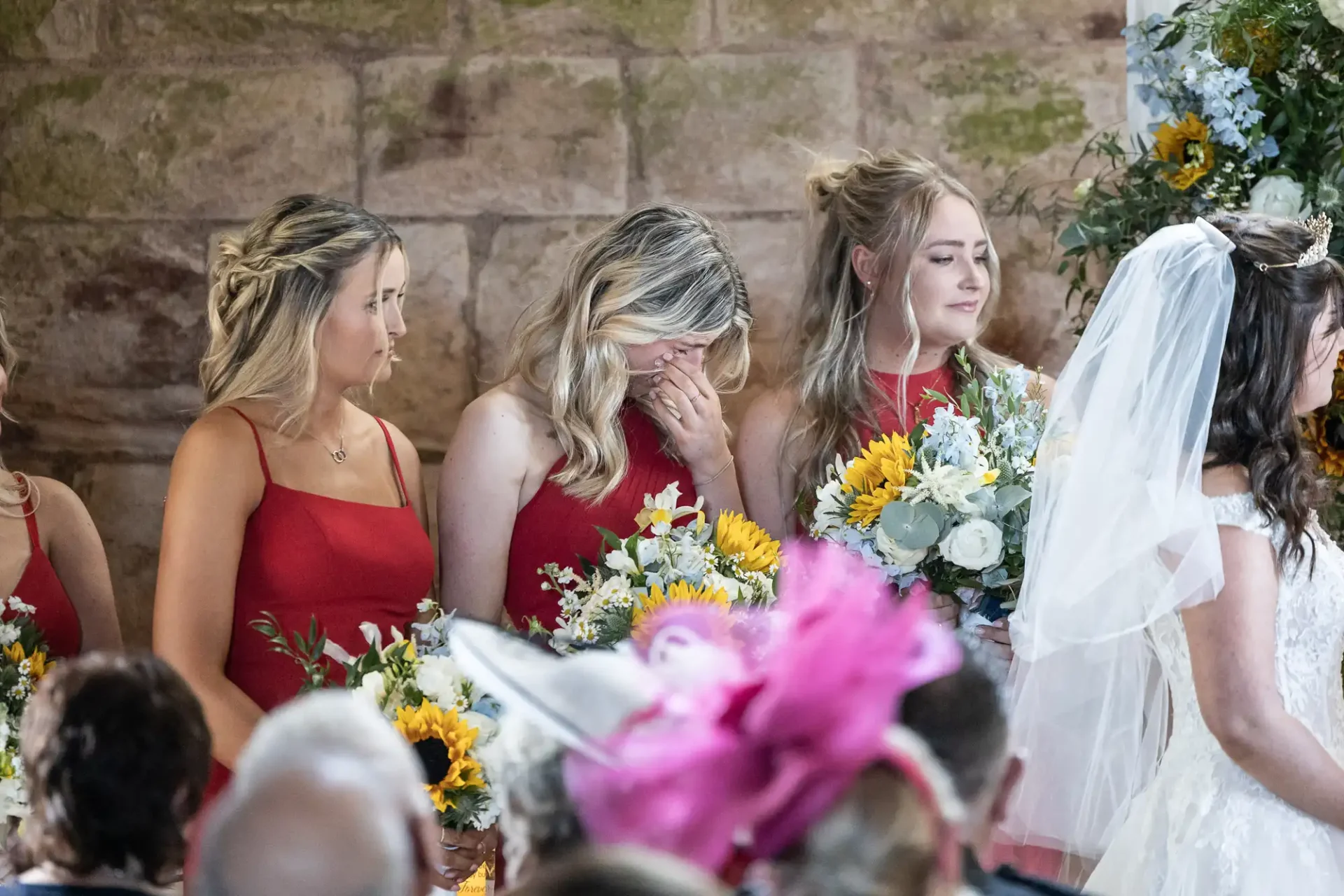 A bride stands with four bridesmaids in red dresses holding flowers, including sunflowers. One bridesmaid uses her hand to cover her face. The setting appears to be an indoor ceremony.