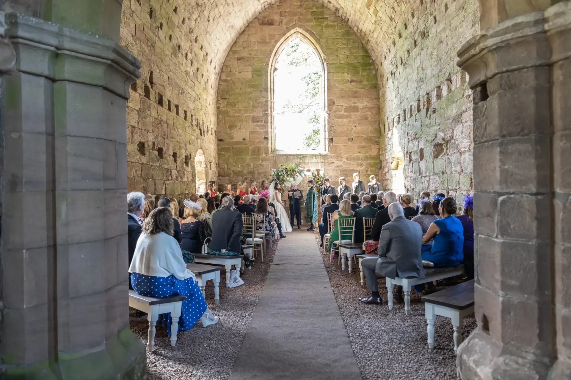 A wedding ceremony inside a stone chapel with guests seated on either side of the aisle, facing the couple and officiant at the front near a large arched window.