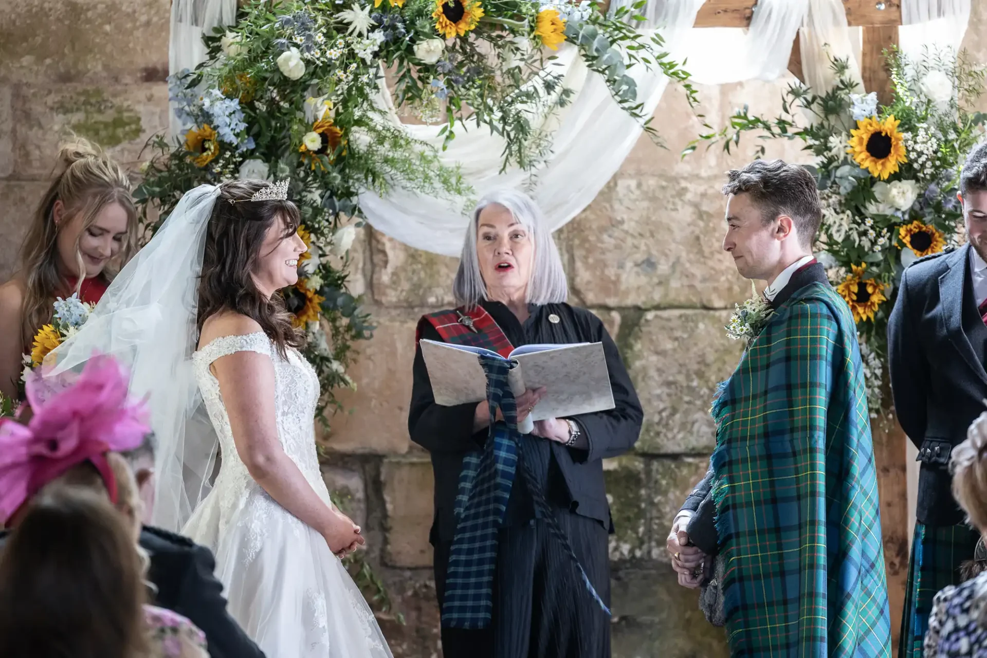 Bride and groom stand before an officiant during a wedding ceremony. They are surrounded by floral arrangements. The groom wears a tartan fabric.