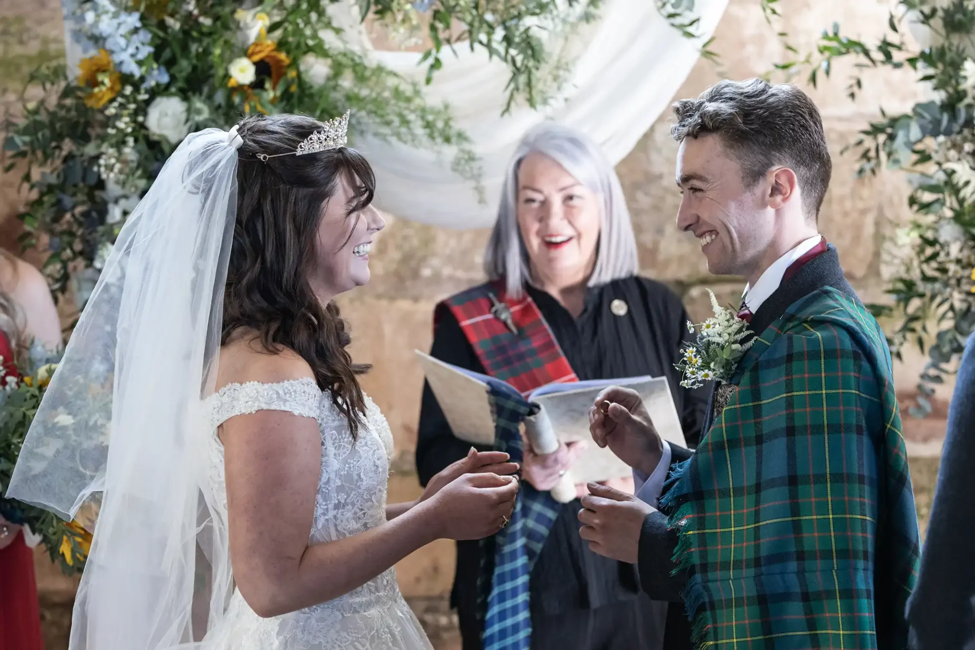 Bride and groom exchange rings during a wedding ceremony, officiated by a woman. The groom wears a tartan shawl, and the bride is in a lace gown with a veil and tiara.