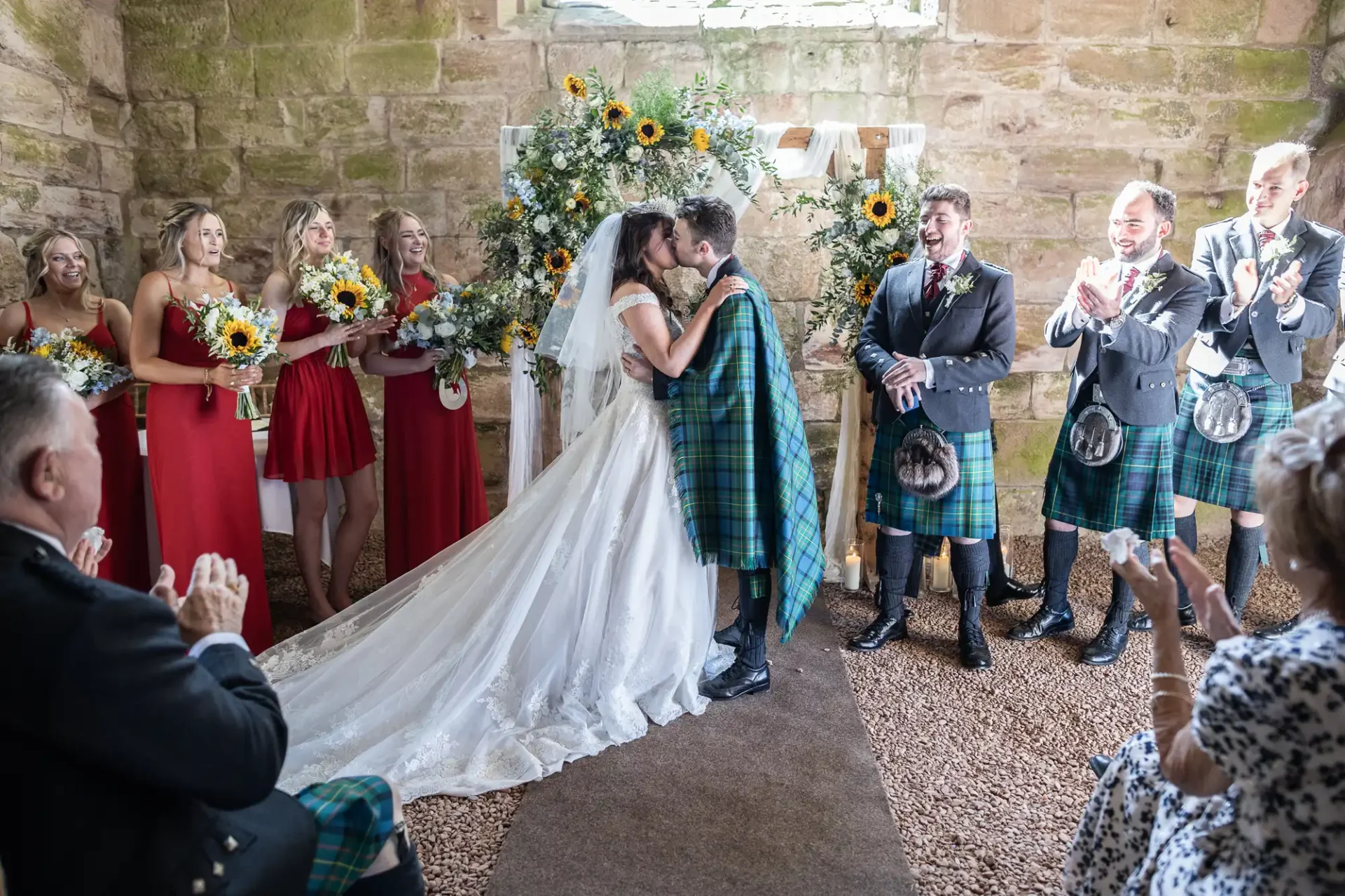 A bride and groom kiss at the altar in a stone-walled venue, surrounded by bridesmaids in red and men in traditional Scottish attire, with attendees clapping.